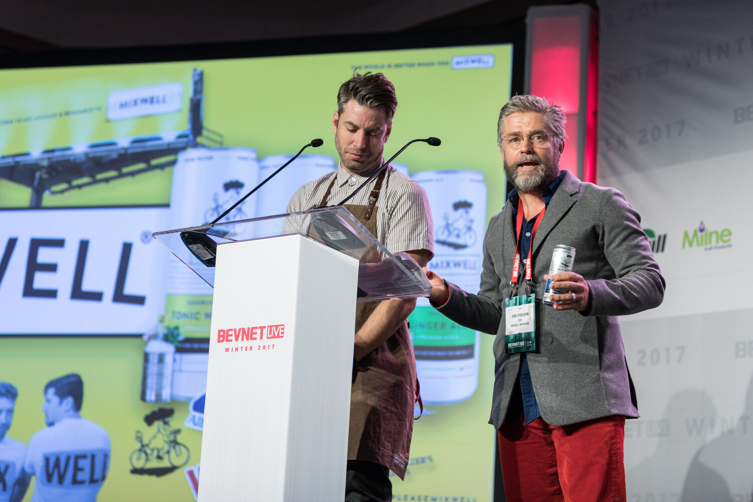 A man in a brown apron looks down at the lectern while a gray-bearded man in a gray blazer and red pants holds a silver can, with a green slide showing WELL cans behind them.