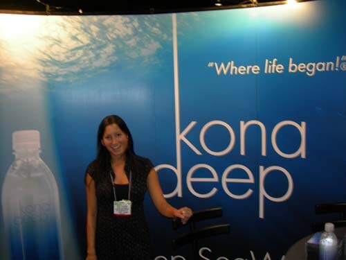 A smiling woman with long dark hair and a badge stands in front of a blue Kona Deep backdrop reading "Where life began!" with a water bottle pictured at left.