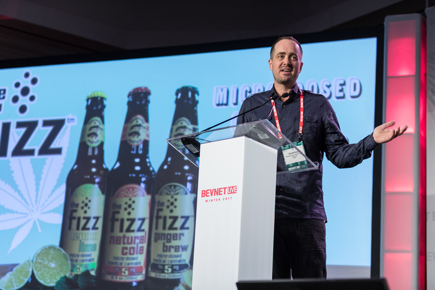 A man in a dark button-up shirt with a headset microphone gestures at a white lectern in front of a slide showing three Fizz bottles labeled natural cola and ginger brew.