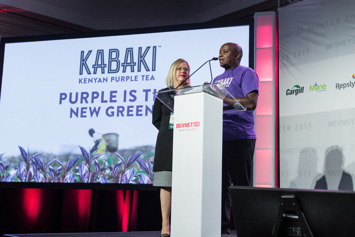 A blonde woman in a black blazer and a man in a purple Kabaki T-shirt stand at a white lectern in front of a slide reading Kabaki Kenyan Purple Tea, Purple is the new green.