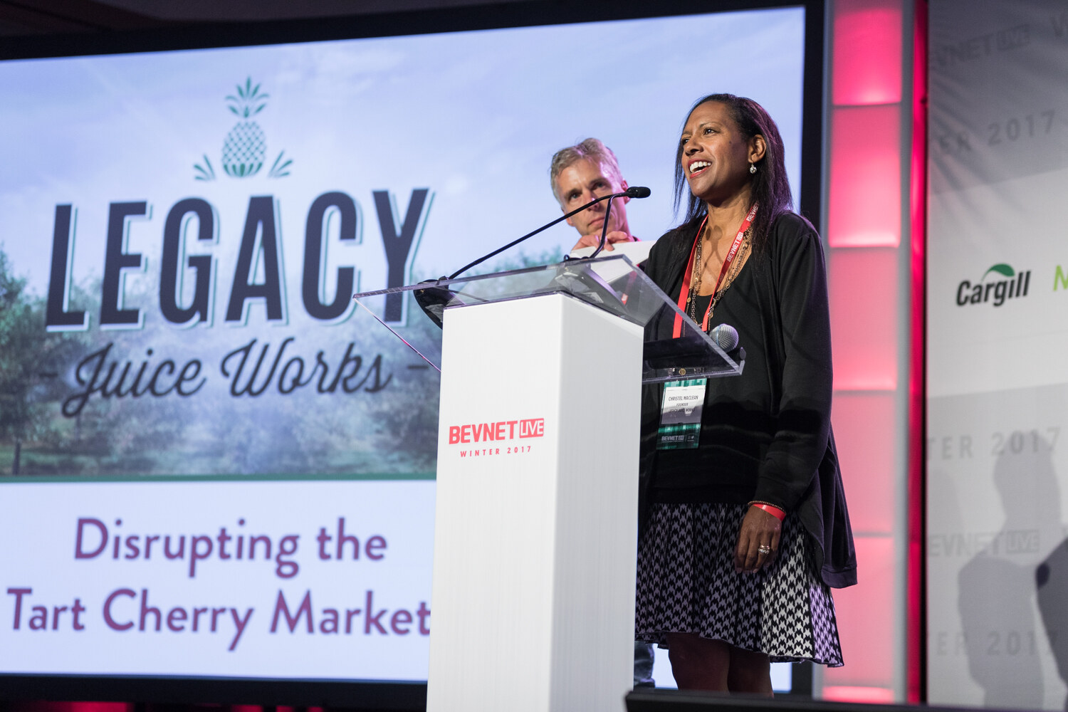 A smiling woman in a black cardigan and patterned skirt speaks at a white lectern in front of a slide reading Legacy Juice Works, Disrupting the Tart Cherry Market, with a gray-haired man behind her.