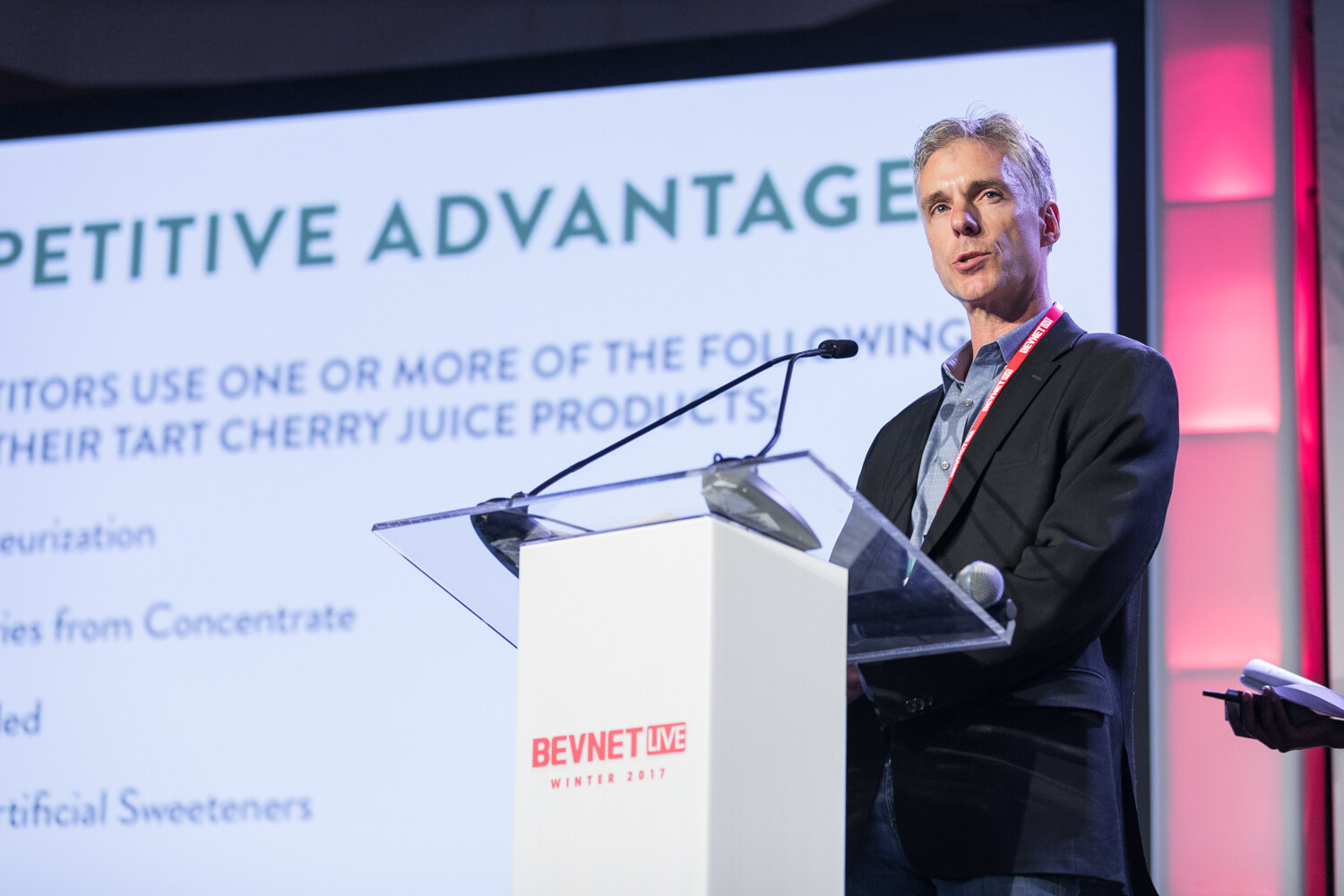 A gray-haired man in a dark blazer and red lanyard speaks at a white lectern in front of a slide headed Competitive Advantage that lists tart cherry juice processing claims.