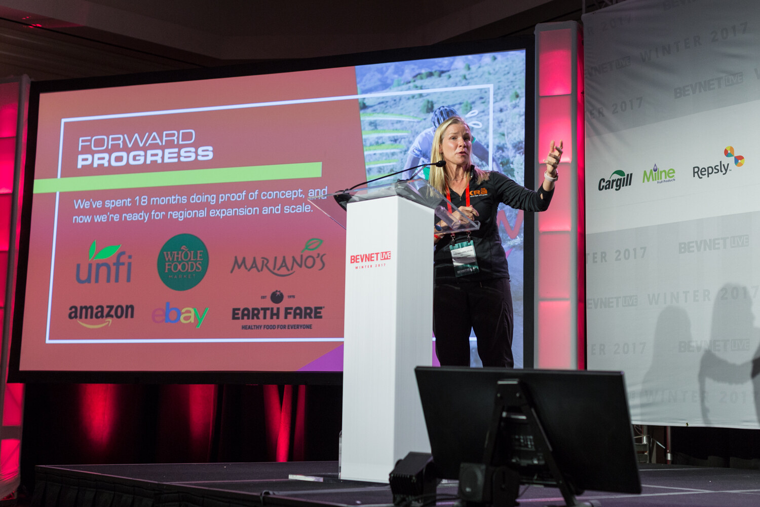 A blonde woman in a black top stands at a white lectern beside a slide titled Forward Progress with the logos of unfi, Whole Foods Market, Mariano's, Amazon, eBay and Earth Fare.