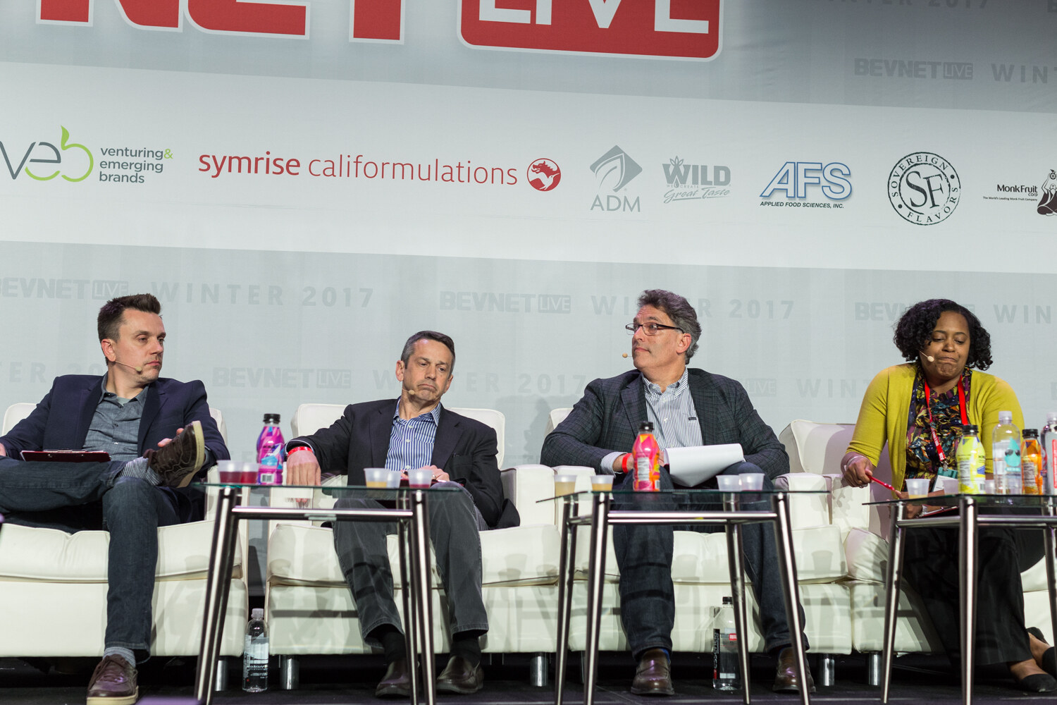 Four judges in blazers and a yellow cardigan sit on white sofas behind glass tables holding bottles and cups under a BevNET Live sponsor banner.