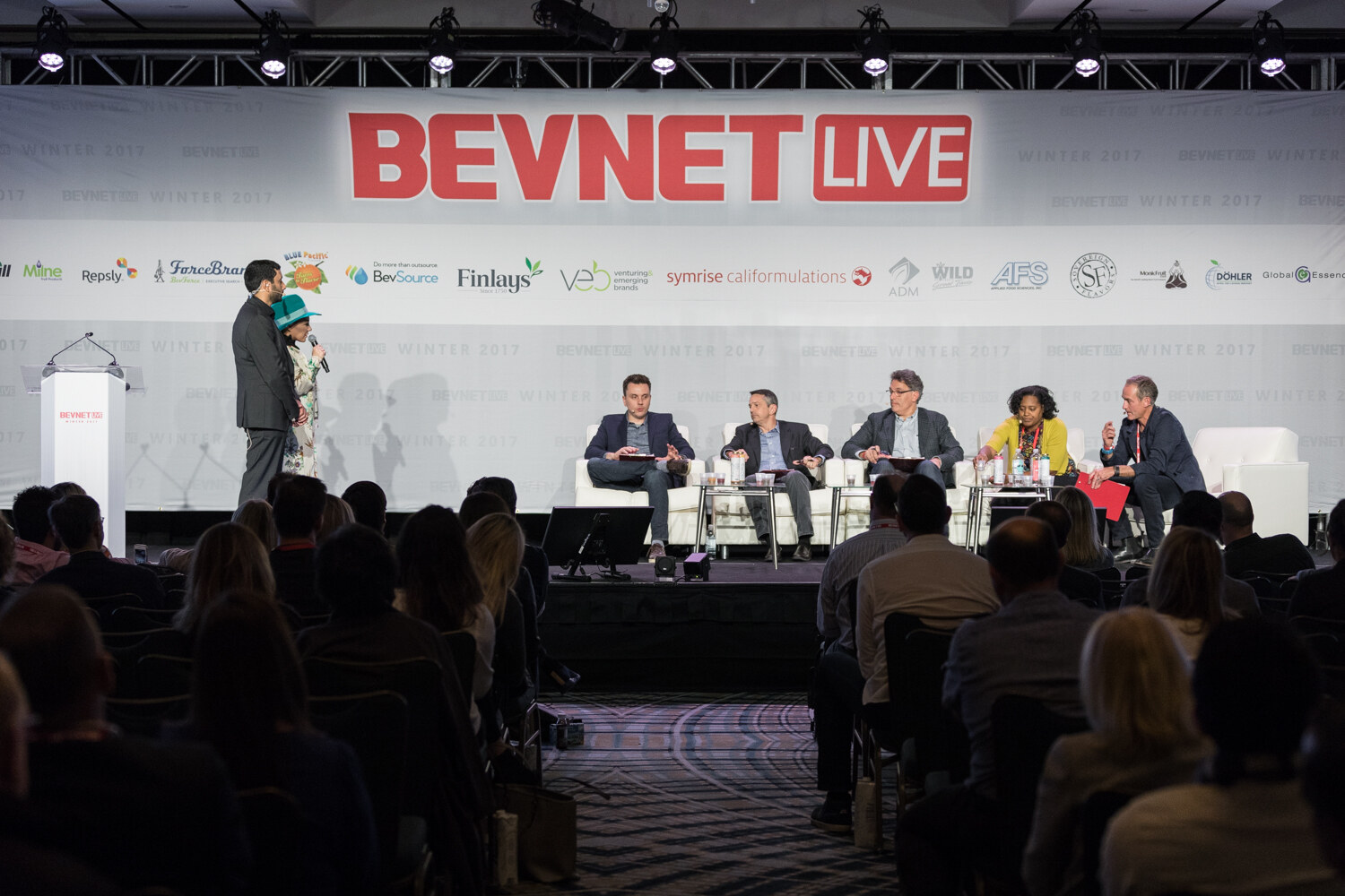 Wide view over the audience of a stage under a BevNET Live banner, with a standing man and a woman in a teal hat at left and five judges seated on white sofas at right.