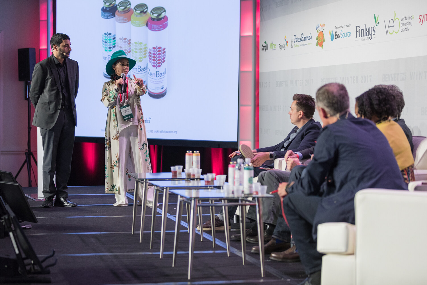 A woman in a teal hat and a floral duster coat holds a microphone in front of a screen showing ZenBasil bottles, with a man in a gray suit beside her and judges seated on sofas at right.