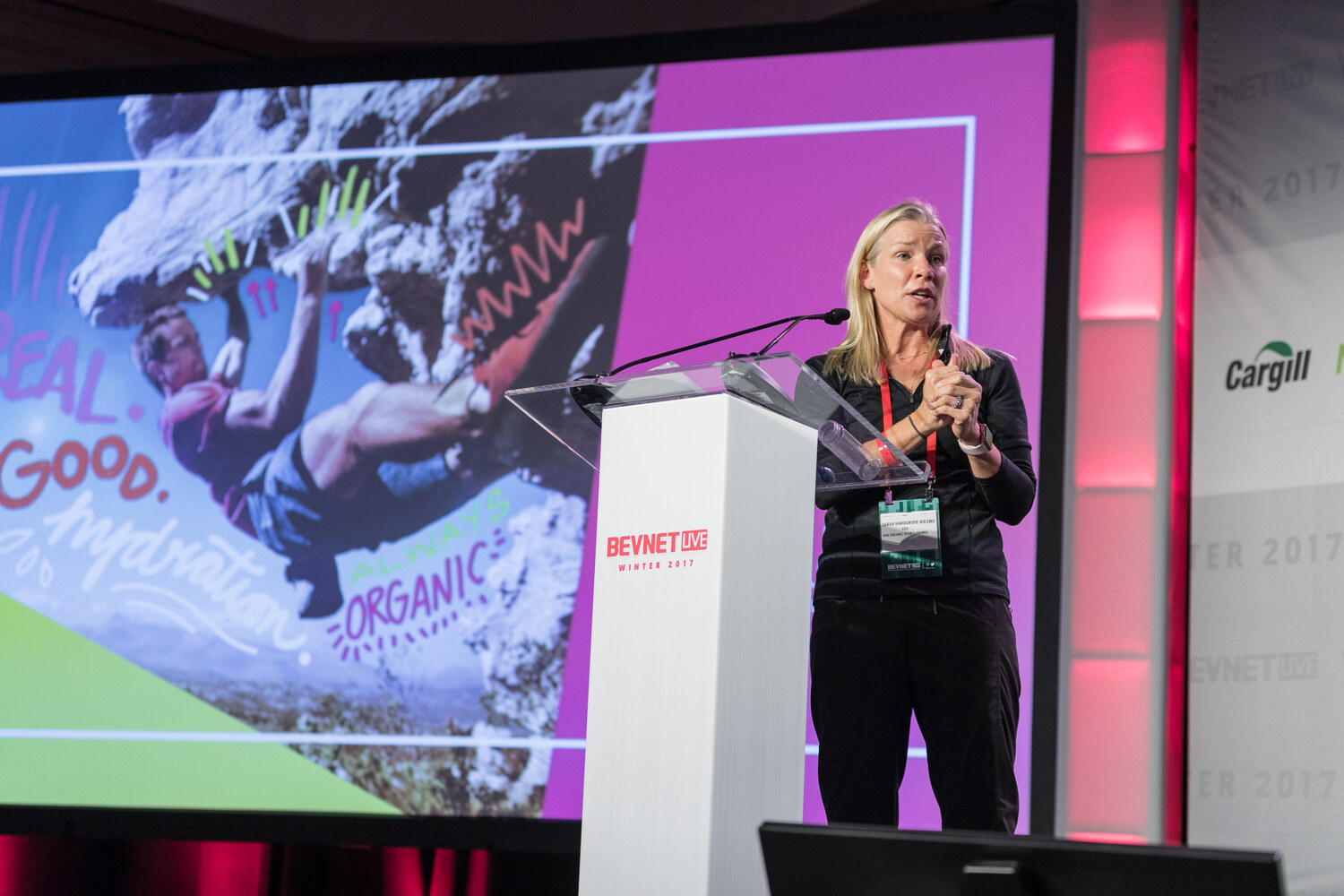 A blonde woman in a black outfit speaks at a white lectern in front of a slide of a rock climber with handwritten text reading Real, Good, Hydration, Always Organic.