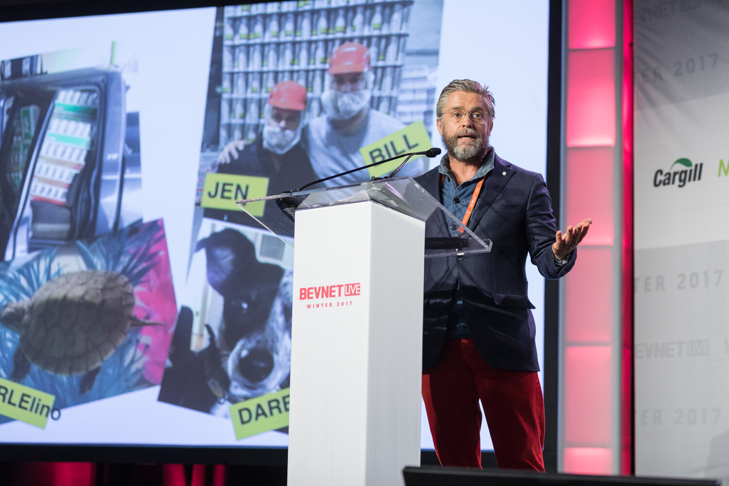 A gray-bearded man in a navy blazer and red trousers opens one hand at a white lectern in front of a slide collage of workers in hairnets, a dog and a turtle with handwritten name labels.