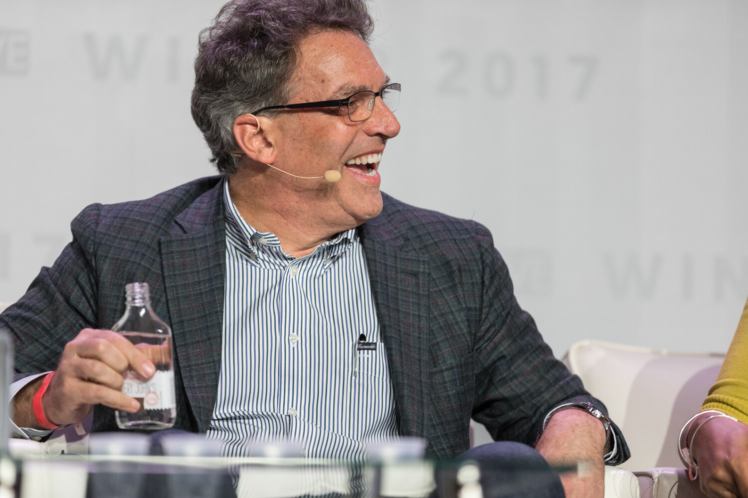 A man in glasses, a plaid blazer and a striped shirt laughs while holding a small glass bottle, with a headset microphone and glass table in front of him.