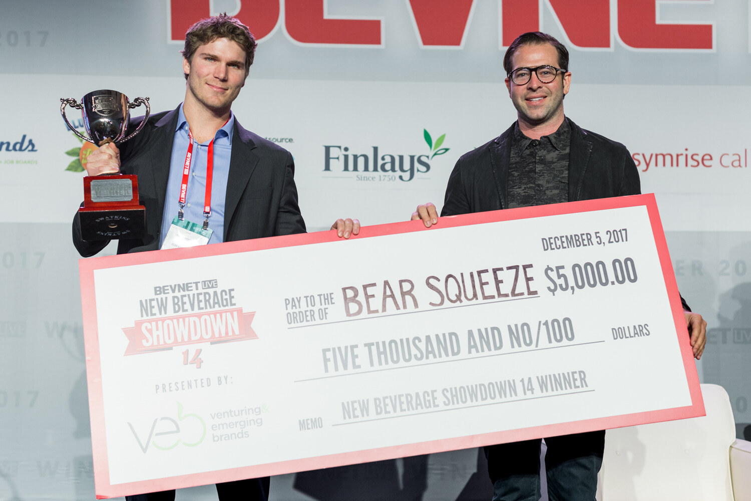 A man holding a silver trophy cup and a man in glasses hold a large check reading Pay to the order of Bear Squeeze, $5,000.00, New Beverage Showdown 14 winner, presented by VEB.