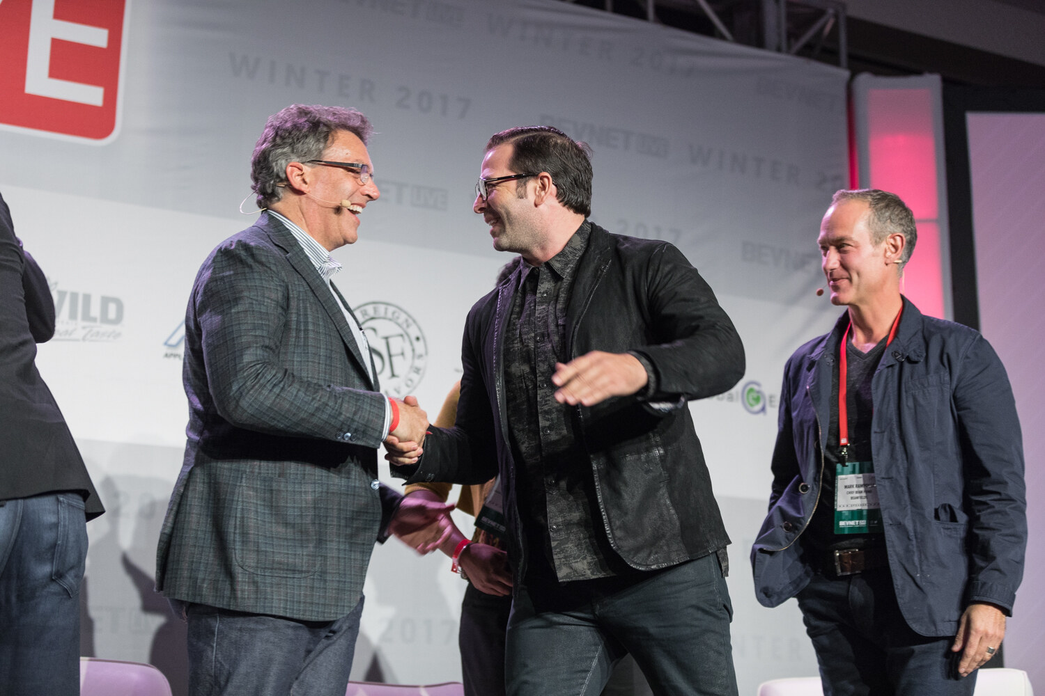 A man in a plaid blazer shakes hands with a smiling man in a black jacket and glasses on a stage while a third man in a navy jacket stands beside them in front of the sponsor backdrop.