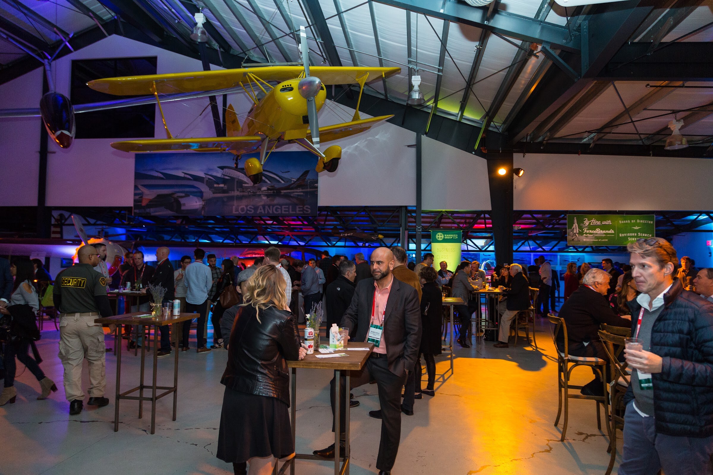 A yellow vintage biplane hangs from the rafters of a hangar above guests standing at high tables, with a green Fly Hire with ForceBrands banner on the far wall.