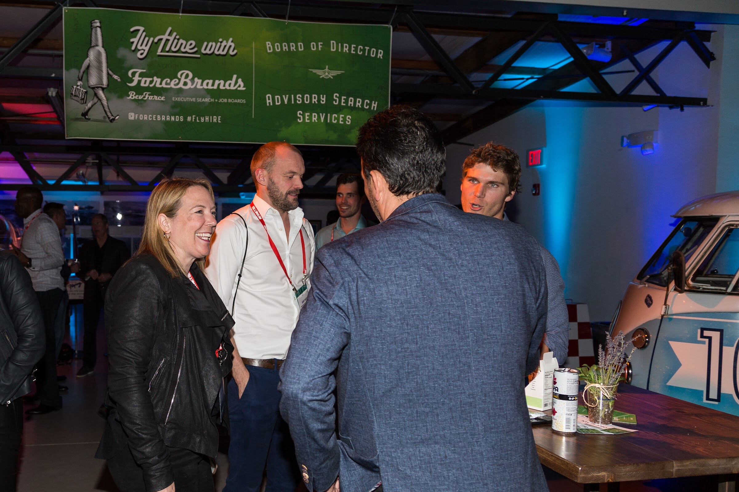A woman in a black leather jacket laughs while talking with three men beneath a green Fly Hire with ForceBrands banner, with a vintage camper van at right.