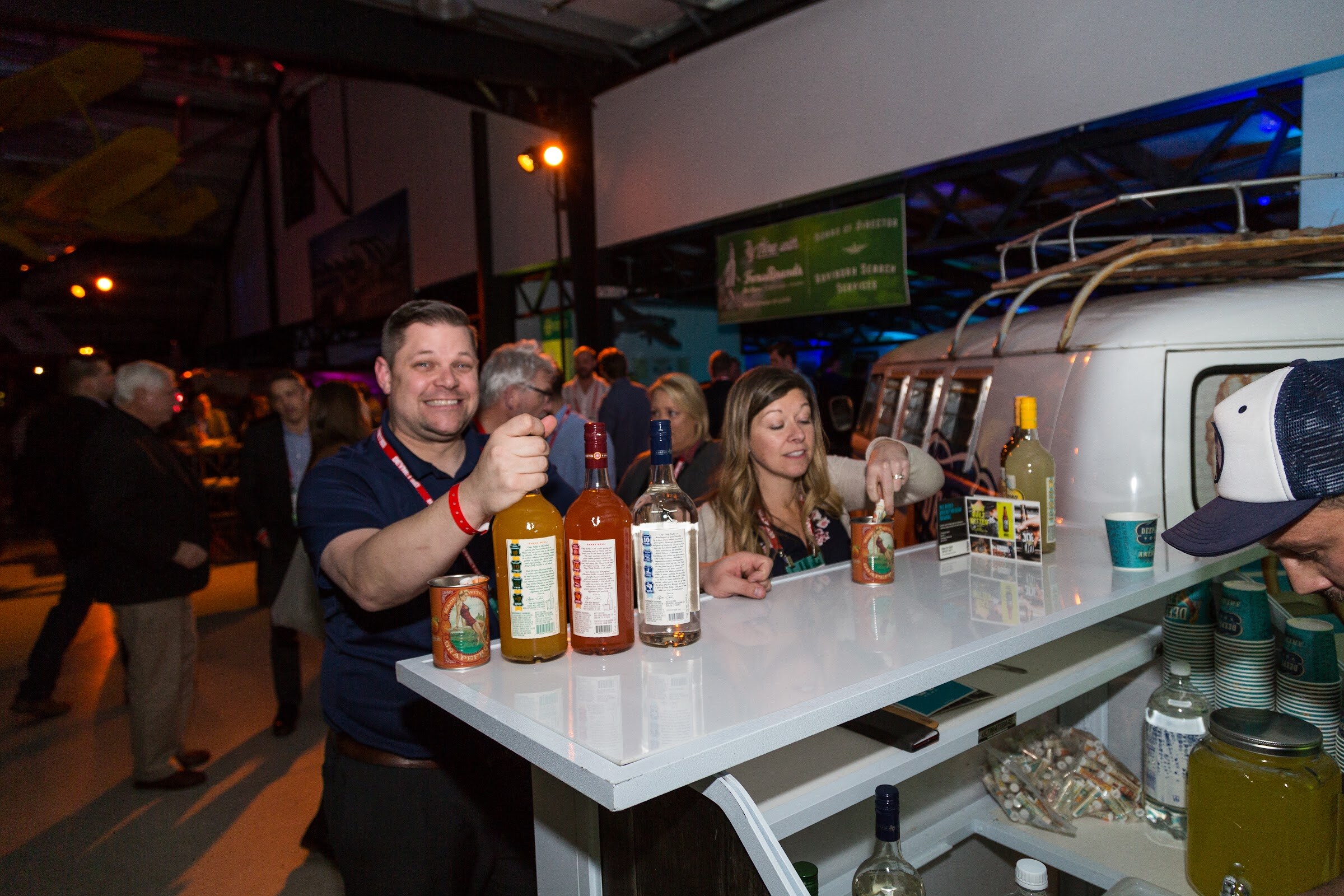 A smiling man in a navy polo holds up a bottle among several bottles on a white bar counter built into a vintage VW van, with a woman beside him and paper cups on a shelf.