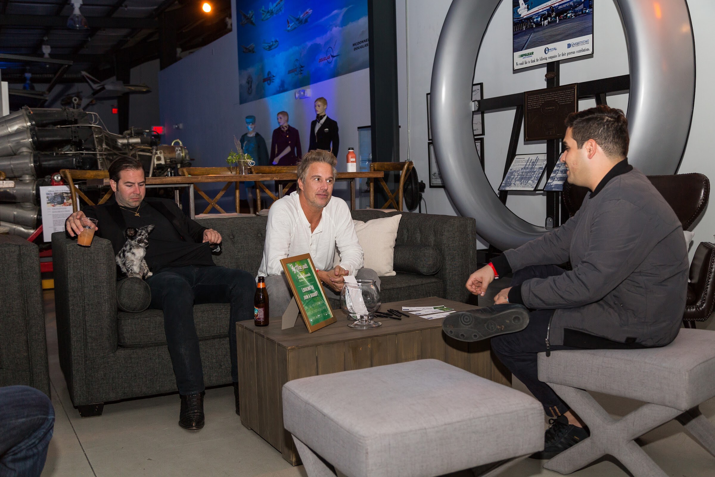Three men converse on gray sofas and a cushioned stool around a wooden coffee table with a beer, in front of a large turbine ring and mannequins in pilot uniforms.
