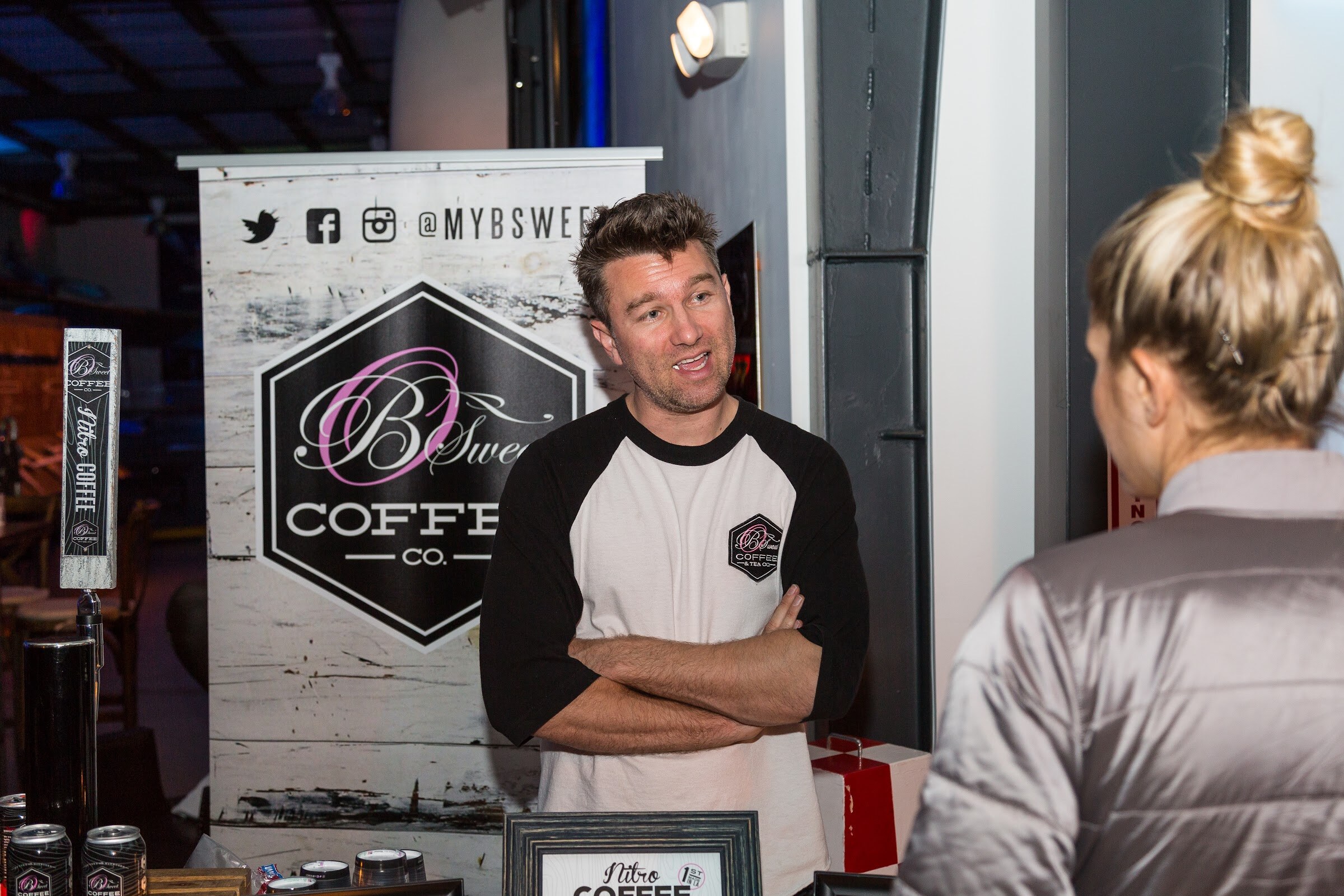 A man in a black-and-white baseball tee with folded arms talks with a woman in front of a B Sweet Coffee Co. banner, with nitro coffee cans and a tap handle nearby.