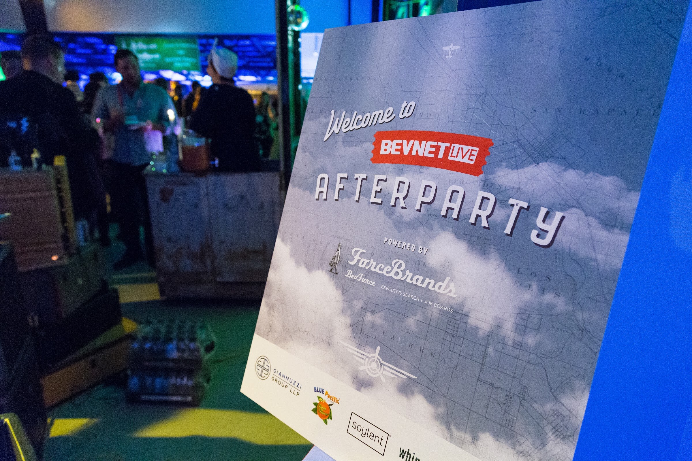 An easel sign reading Welcome to BevNET Live Afterparty, powered by ForceBrands, with sponsor logos for Giannuzzi Group LLP, Blue Pacific and Soylent, in front of guests at a bar in a dim hangar.