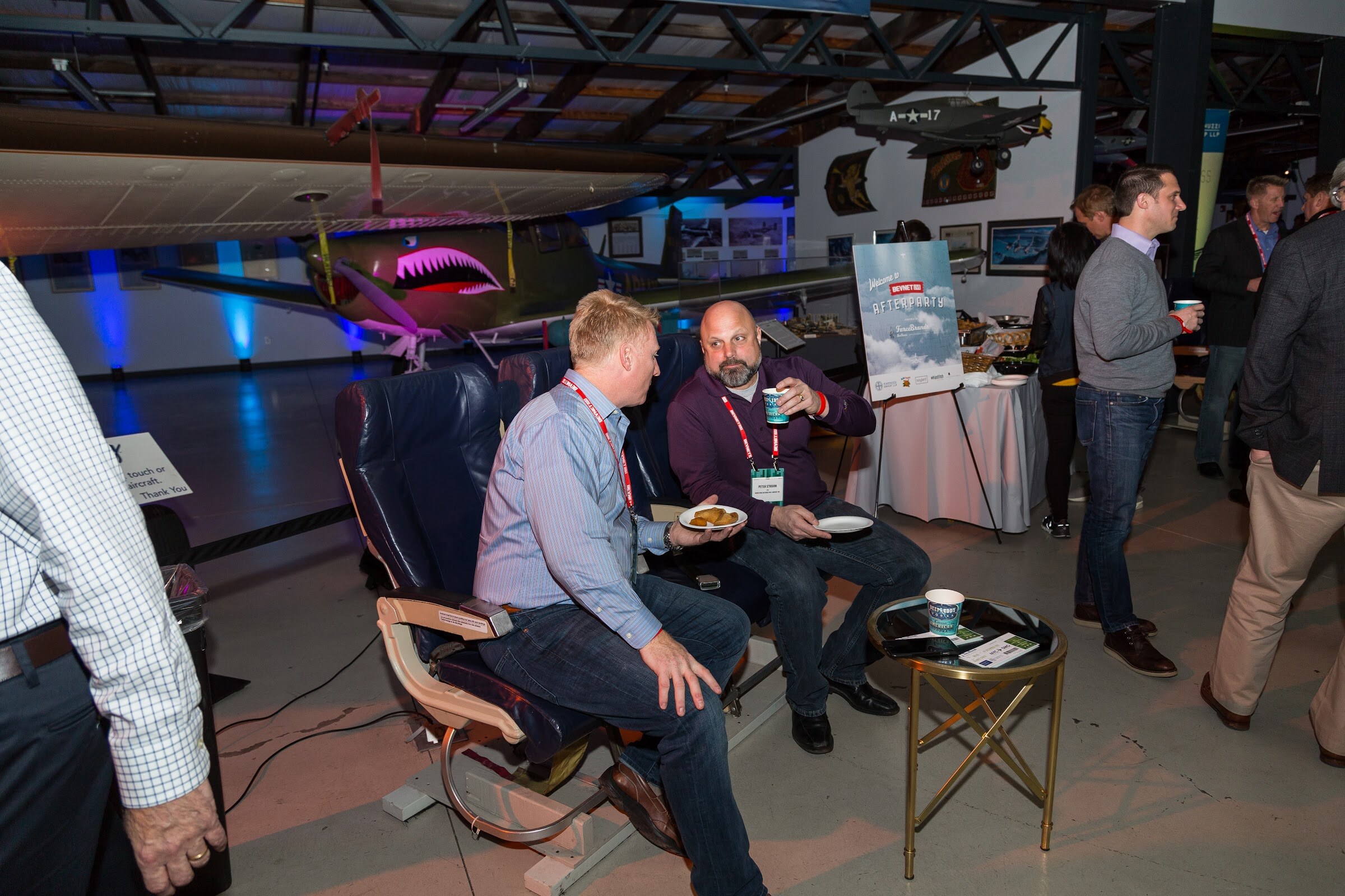 Two men with red lanyards sit in navy vintage airline seats holding plates and cups, with a World War II fighter plane painted with a shark mouth behind them and a Welcome to BevNET Live Afterparty sign at right.