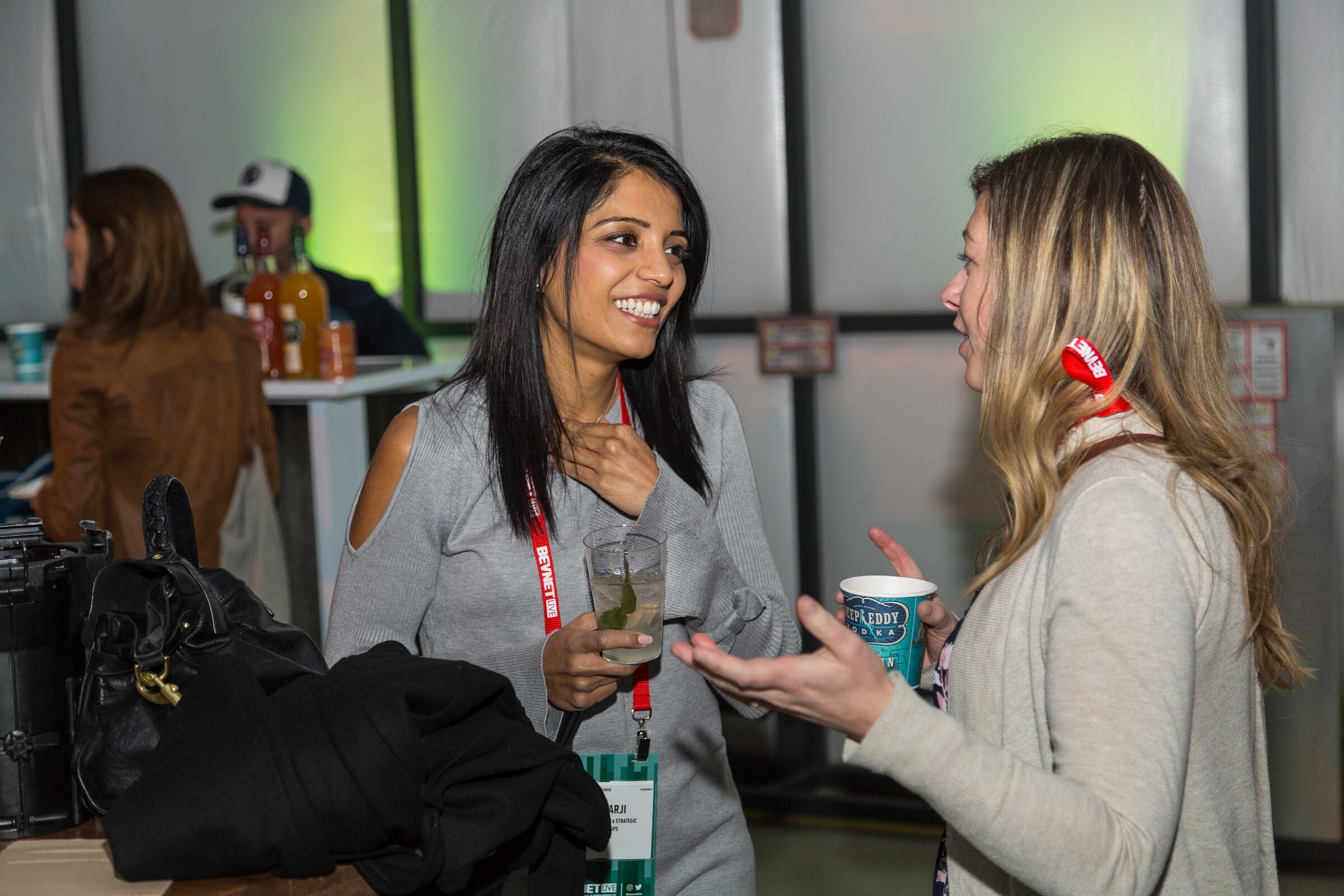A smiling woman with long dark hair in a gray cold-shoulder top holding a drink talks with a blonde woman holding a Deep Eddy cup, against a green-lit wall.