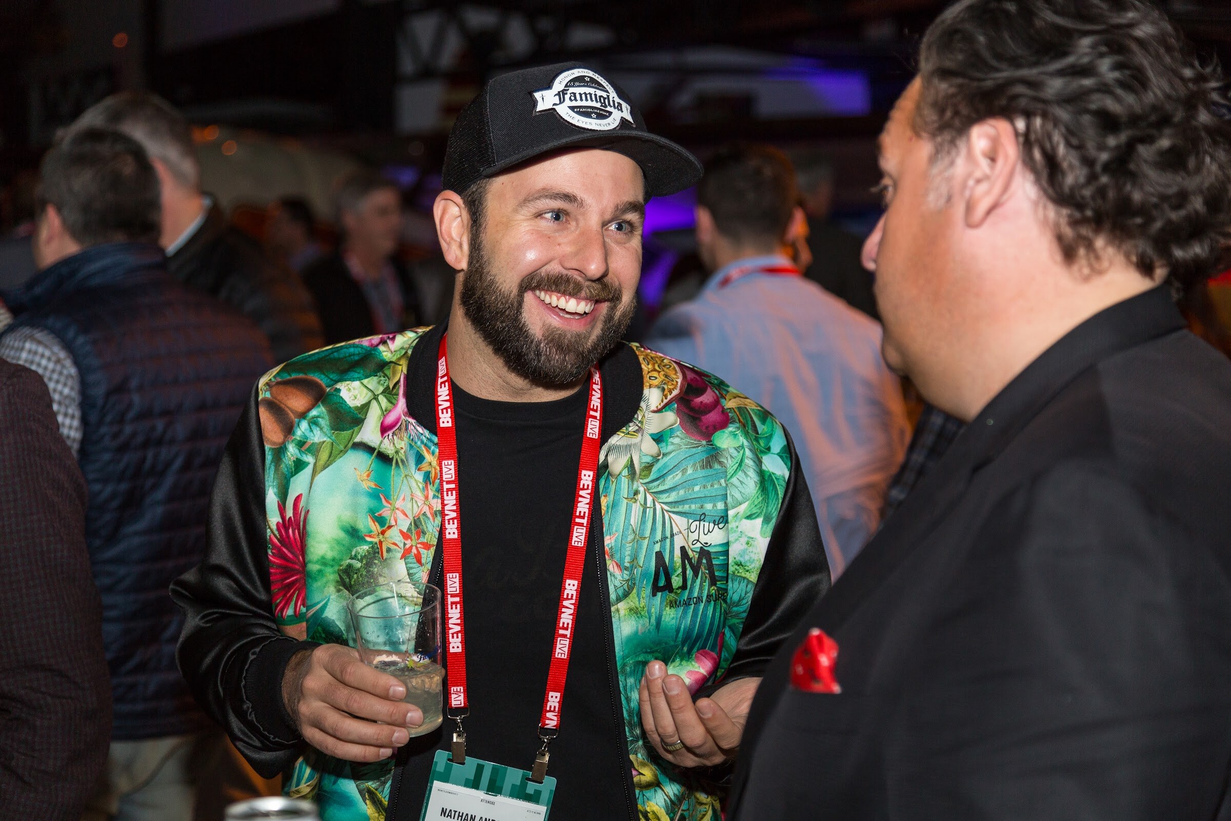 A smiling bearded man in a black cap and a tropical-print bomber jacket with a red BevNET Live lanyard holds a glass while talking with a man in a dark blazer with a red pocket square.