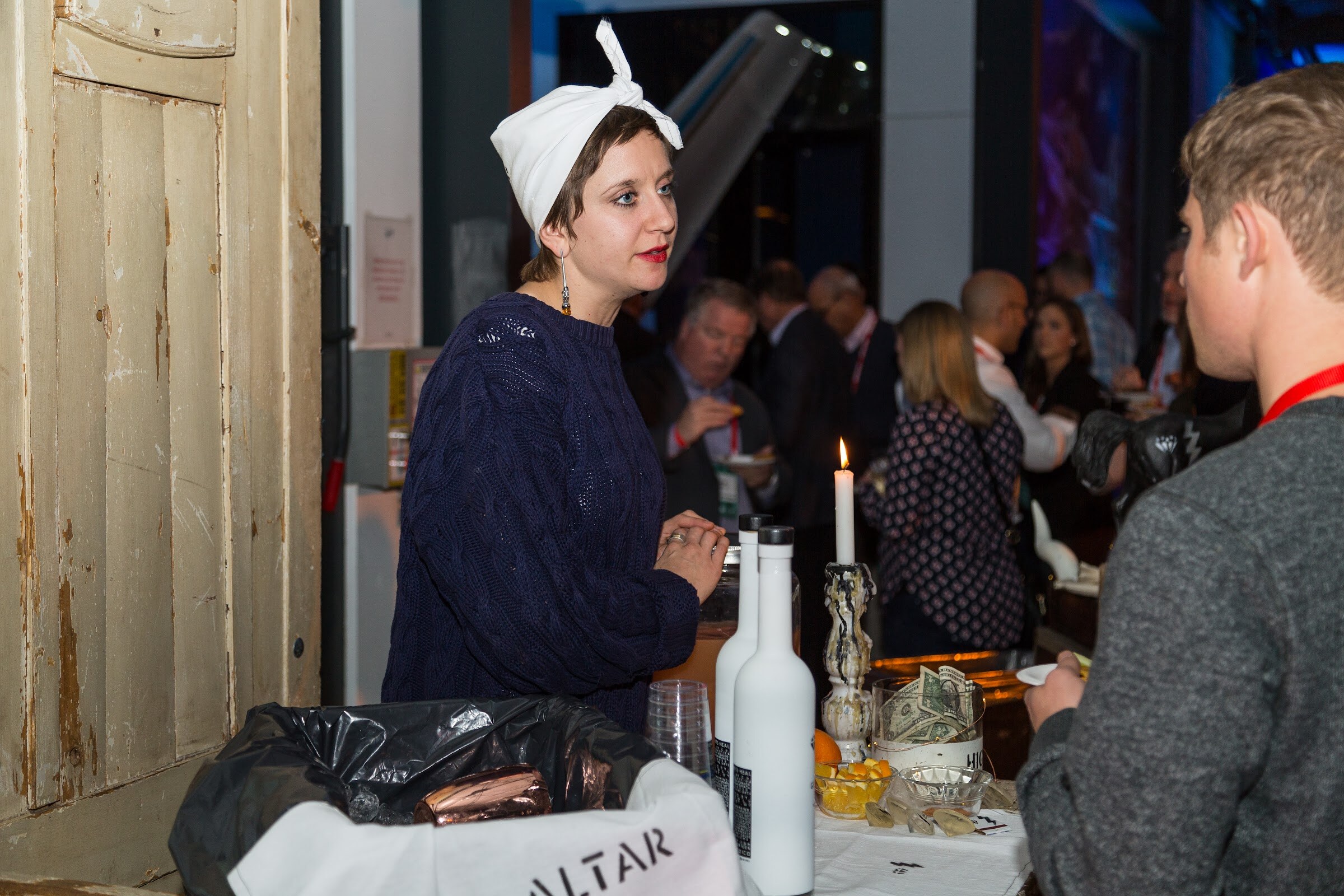 A woman in a white head wrap and navy cable-knit sweater stands behind a bar with white bottles, a lit candle and a bowl of cash, with a bag labeled Altar in the foreground and guests behind her.