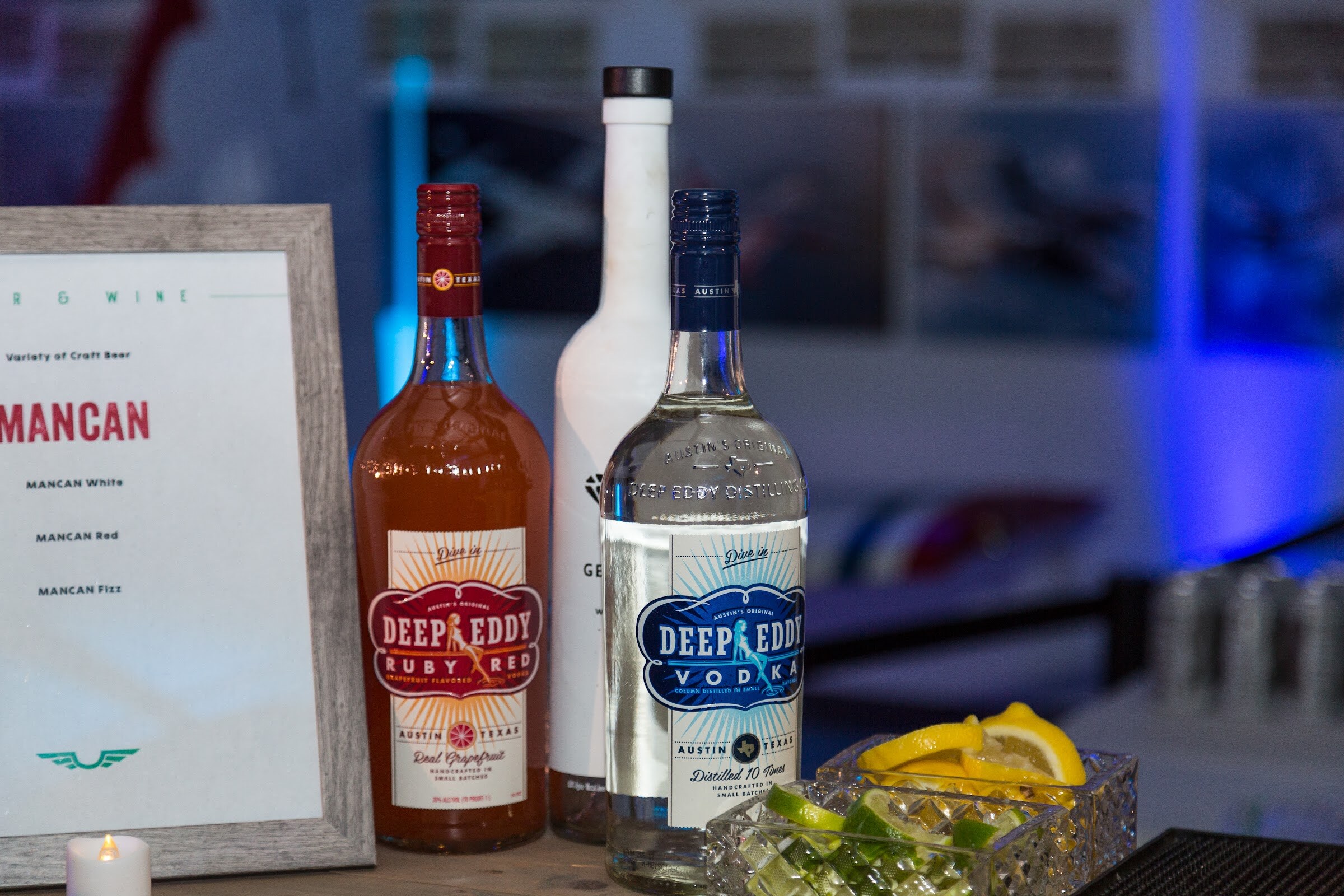 Two bottles labeled Deep Eddy Ruby Red and Deep Eddy Vodka beside a framed menu listing MANCAN White, Red and Fizz, with a bowl of lemon and lime wedges in front.