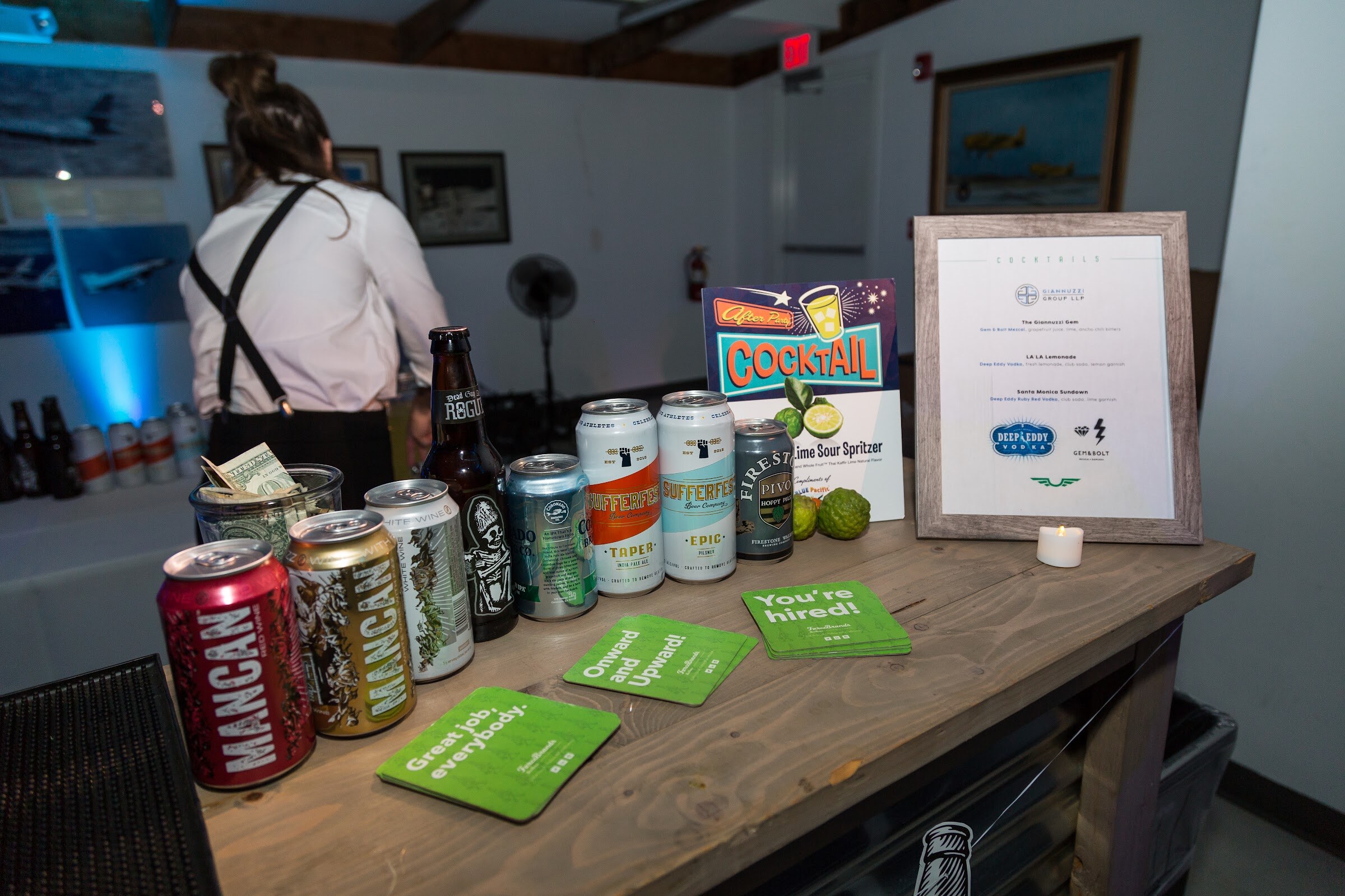 A wooden bar top with cans of MANCAN, Sufferfest and Firestone, green coasters reading Great job, everybody, Onward and Upward and You're hired, and a framed cocktail menu listing Deep Eddy Vodka drinks.