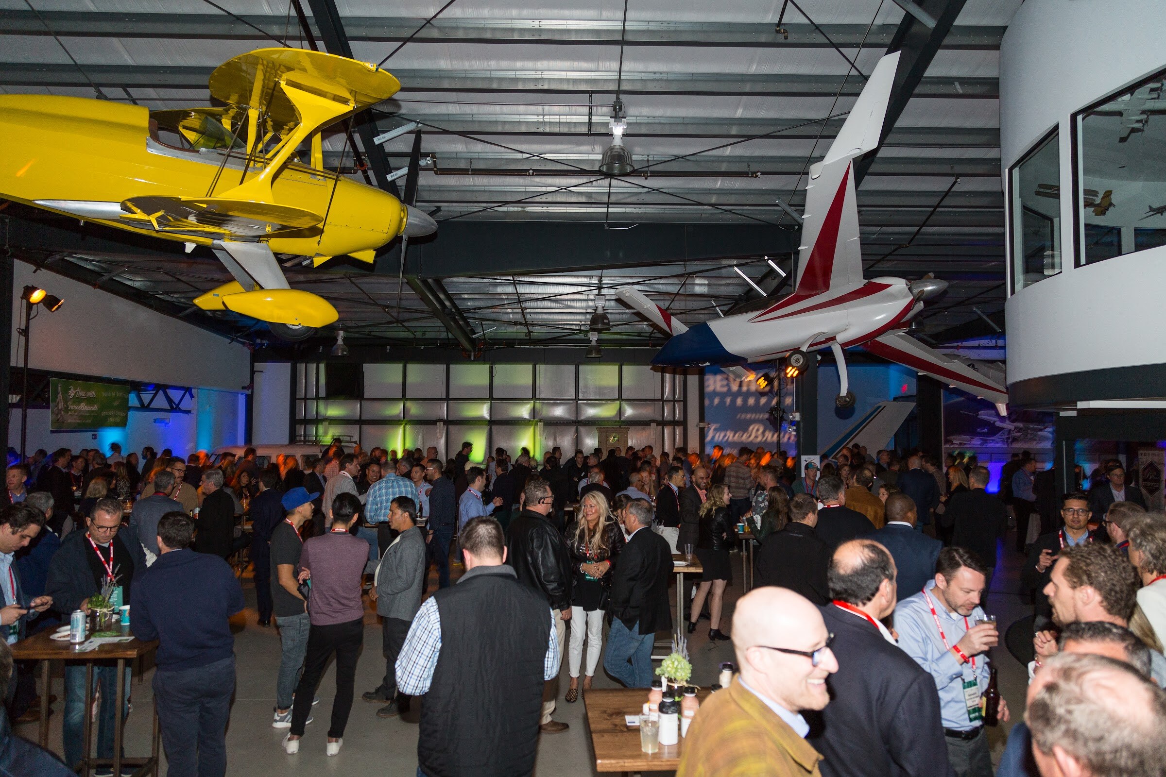 Wide view of a crowded hangar party with a yellow biplane and a red and white plane suspended overhead and guests gathered around wooden high tables.