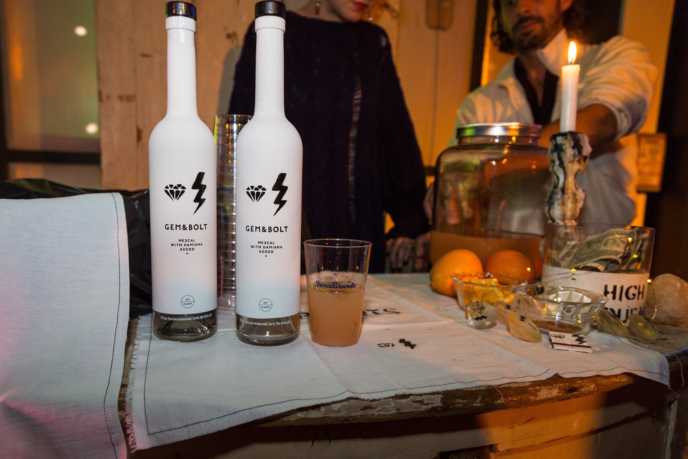 Two white Gem&Bolt mezcal bottles stand on a bar covered with a cloth, next to a ForceBrands cup of punch, oranges, a tip jar and a lit candle, with bar staff behind.