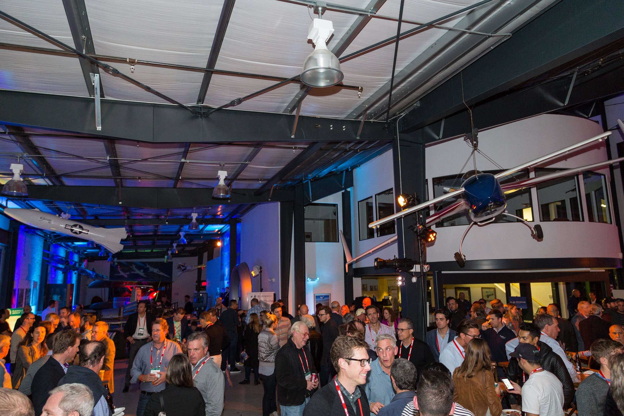 A crowded hangar party with attendees wearing red lanyards talking beneath suspended airplanes under a corrugated ceiling.