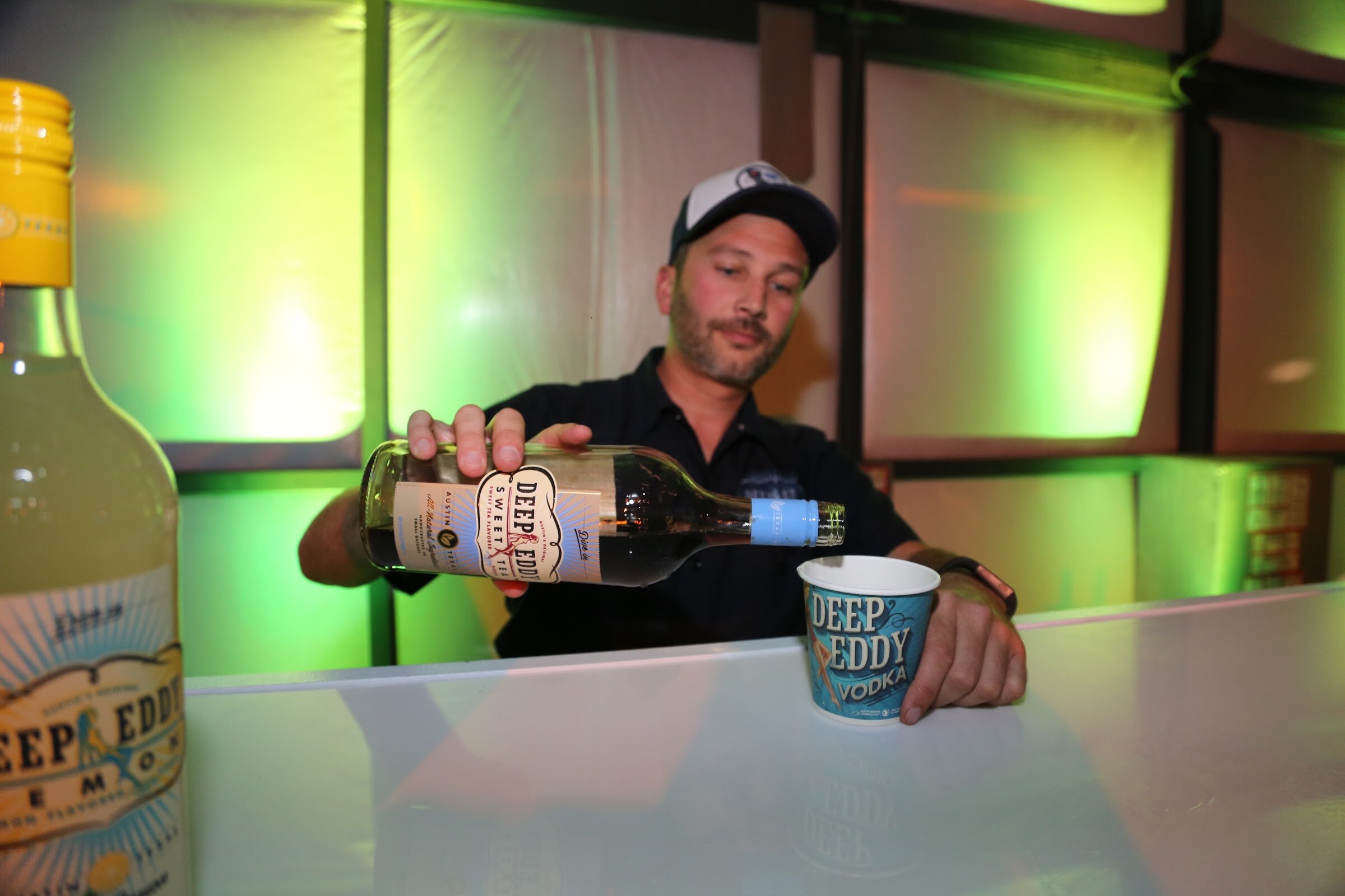 A bearded bartender in a baseball cap pours from a Deep Eddy bottle into a Deep Eddy Vodka cup on a bar lit in green, with a Deep Eddy Lemon bottle at left.