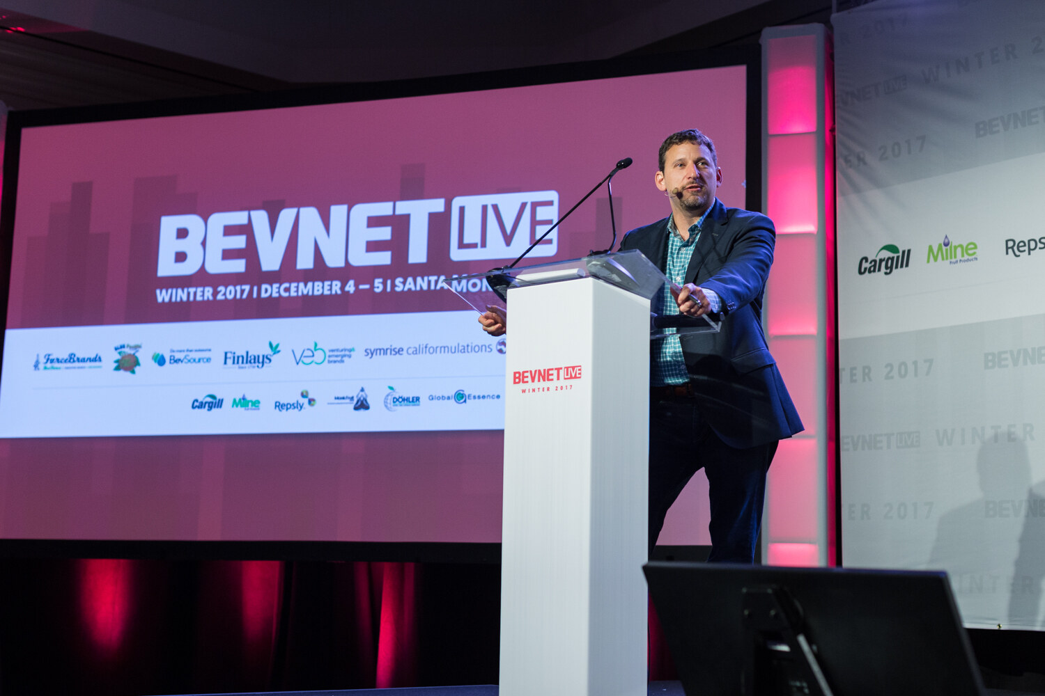 A man in a navy blazer and teal checked shirt speaks at a white lectern in front of a large screen reading BevNET Live Winter 2017, December 4-5, Santa Monica, with sponsor logos.