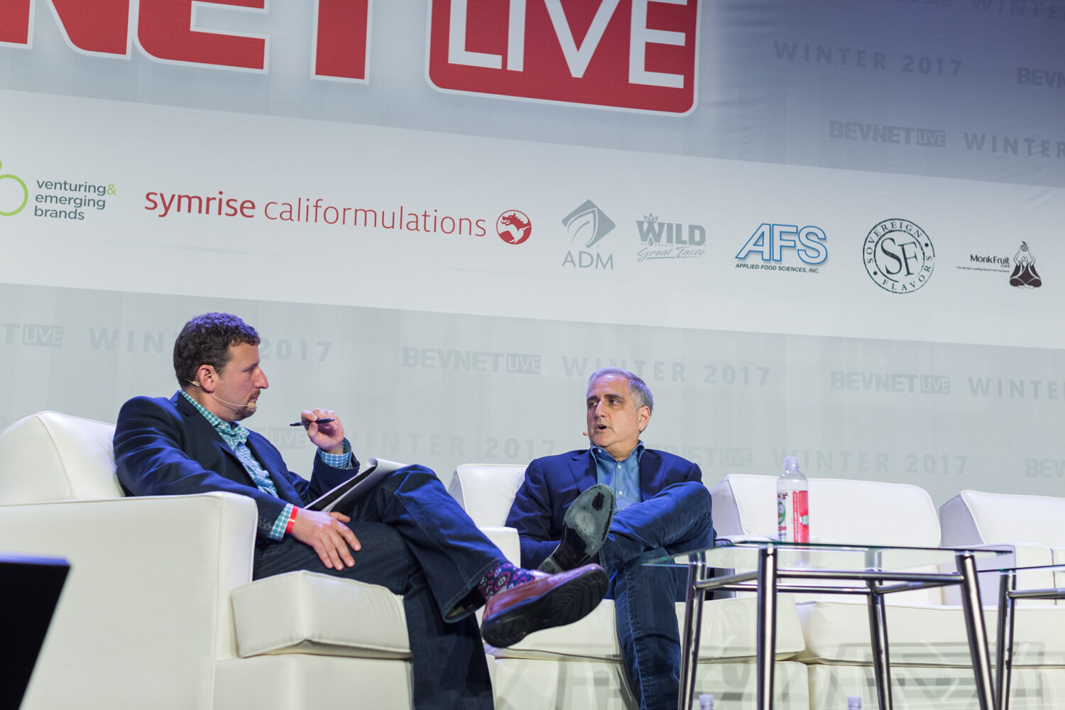 Two men in blazers sit on white sofas on stage, one holding a pen and tablet, with a BevNET Live banner and sponsor logos above them.