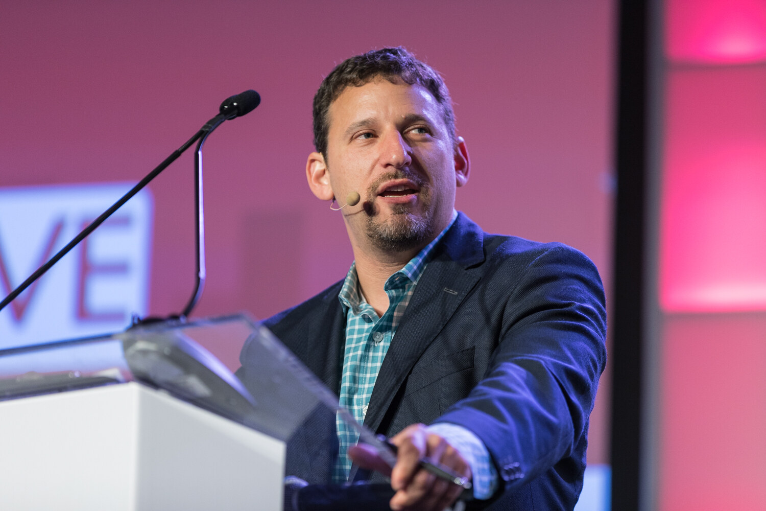 Close-up of a bearded man in a headset microphone and navy blazer speaking at a clear lectern against a pink backdrop.