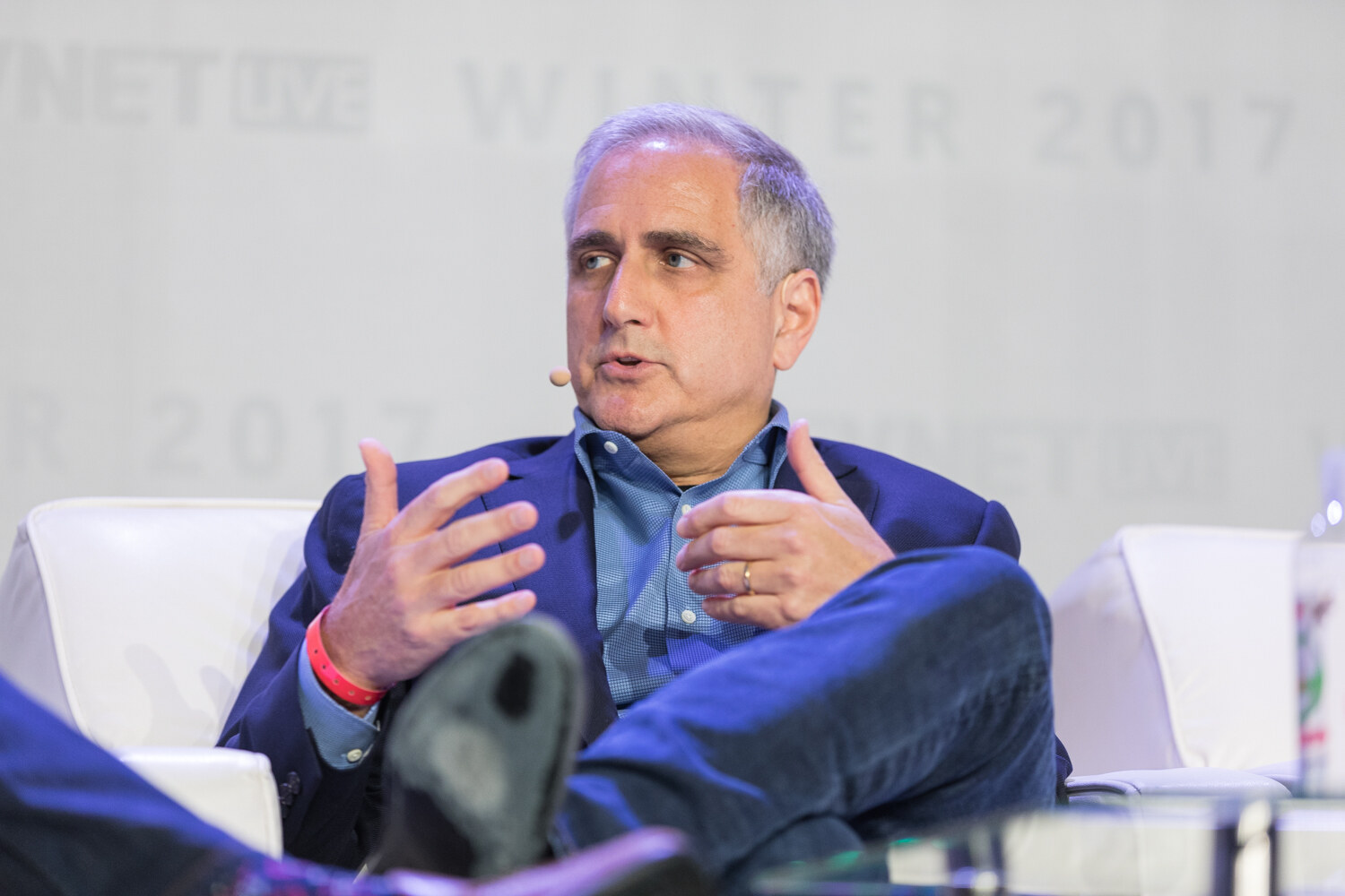 A gray-haired man in a blue blazer gestures with both hands while speaking, seated on a white sofa with a headset microphone.