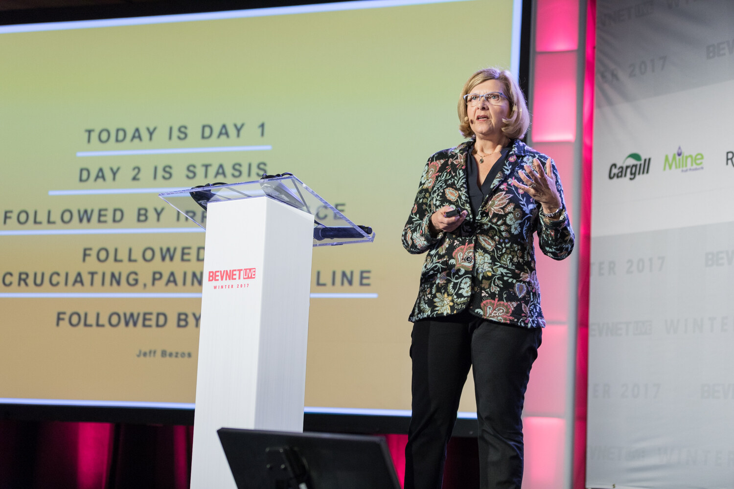 A woman with short blonde hair and glasses in a floral blazer stands next to a white lectern, with a slide behind her that begins Today is Day 1, Day 2 is stasis and is credited to Jeff Bezos.