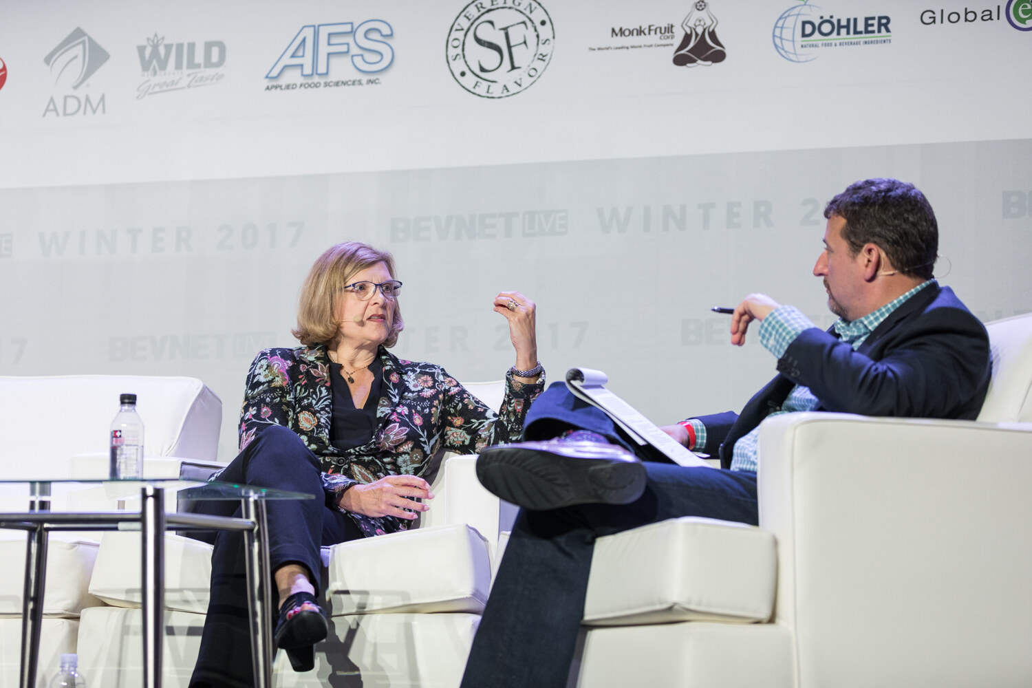 A woman in a floral jacket gestures while seated across from a man with a notepad and pen on a stage with white sofas and sponsor logos.