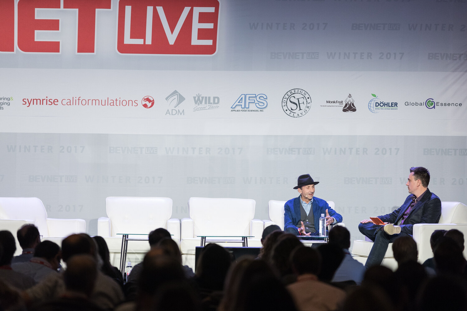 Wide view from the audience of two men talking on a stage with white sofas under a BevNET Live banner and sponsor logos.