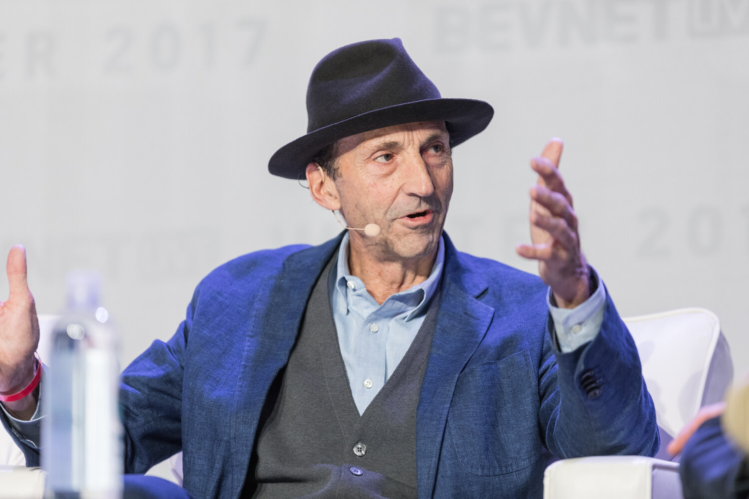 Close-up of an older man in a black fedora, blue blazer and gray vest speaking with a headset microphone and raising one hand.