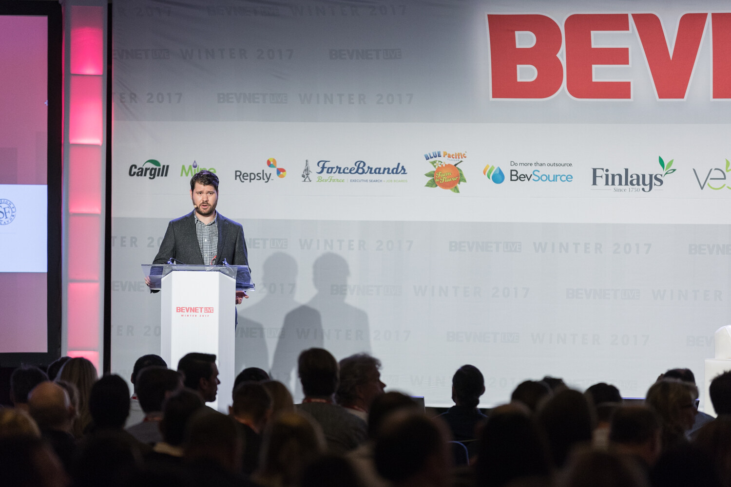 A man at a white lectern in front of a BevNET Live backdrop with sponsor logos, seen over the heads of the audience.