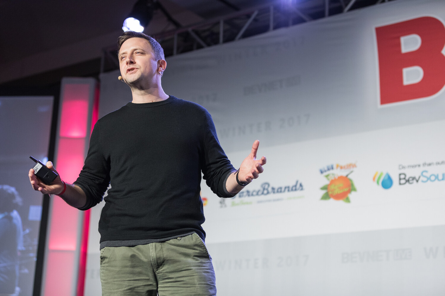 A man in a black sweater and olive pants with a headset microphone holds a slide clicker in one hand and gestures with the other on a stage with sponsor logos behind.