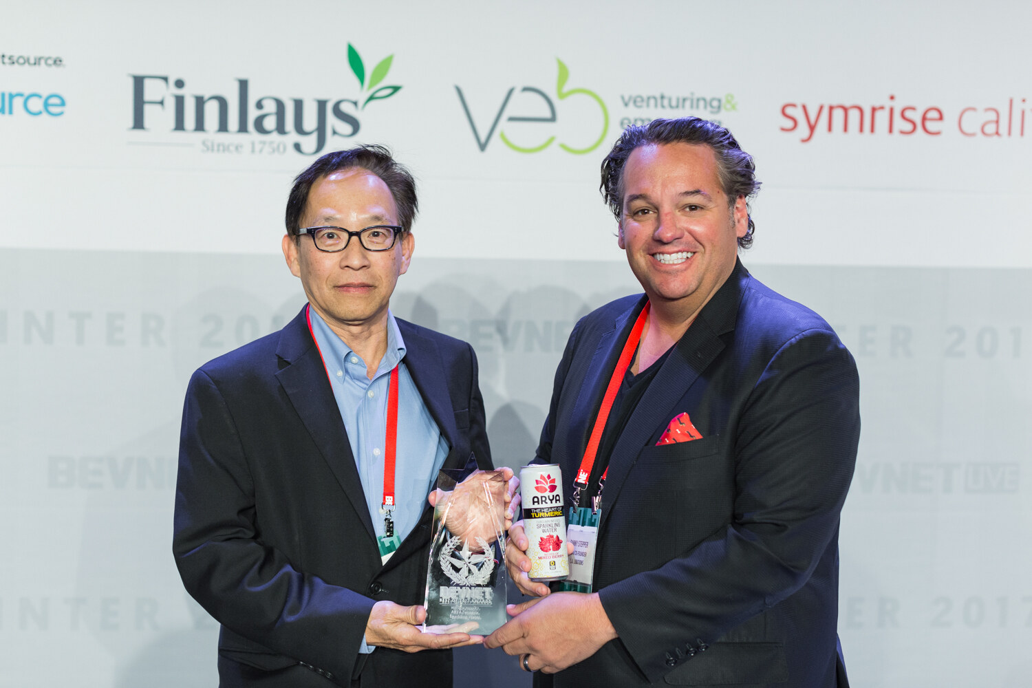 Two men in dark blazers hold a glass award and an Arya sparkling water can in front of a backdrop with Finlays and VEB logos.