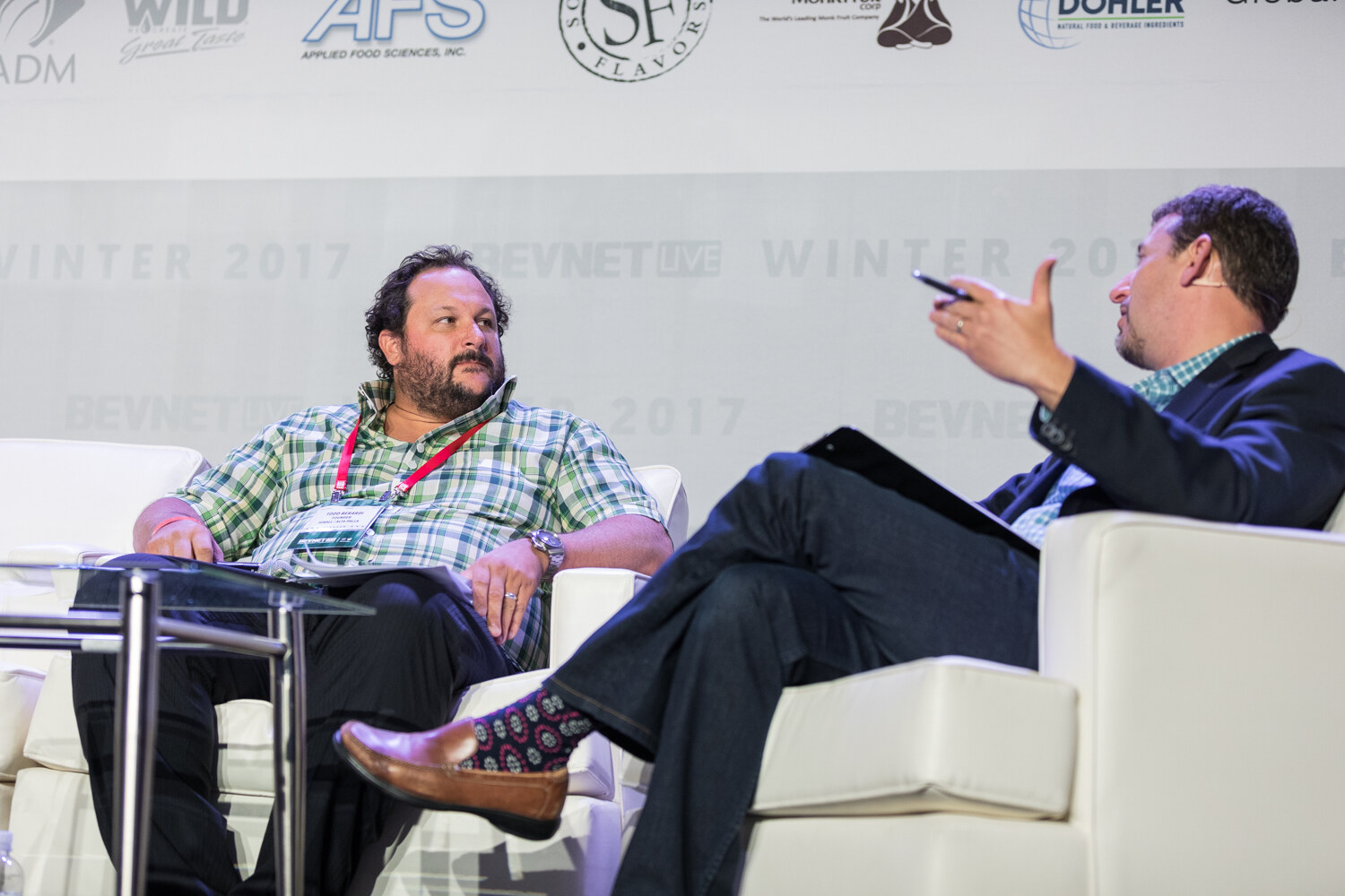 Two men on white sofas on stage, one in a green plaid shirt with a lanyard looking across at the other, who gestures with a pen and clipboard.