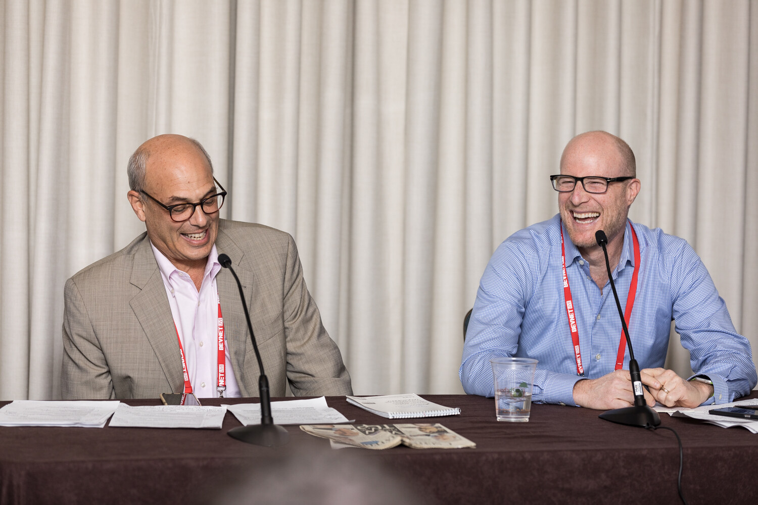 Two bald men with glasses laugh while seated at a table with microphones and papers in front of cream curtains, wearing red BevNET lanyards.