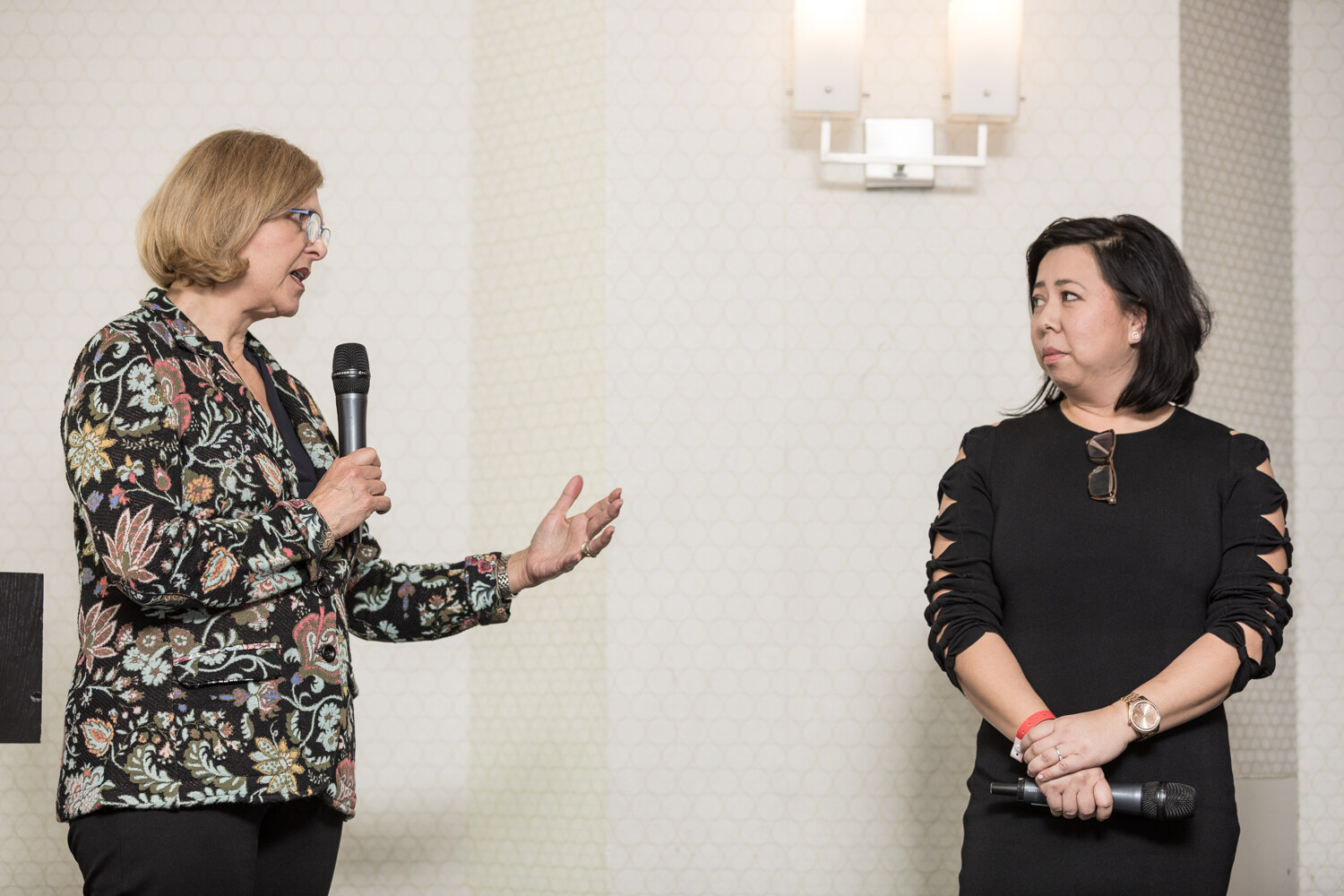 A woman in a floral jacket speaks into a handheld microphone and gestures, while a woman in a black dress with cutout sleeves looks on holding a microphone.