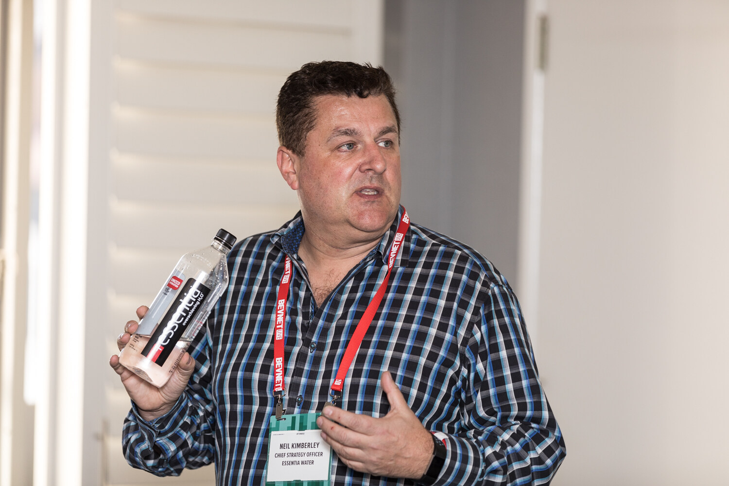 A man in a dark plaid shirt with a red lanyard holds a clear Essentia water bottle in one hand and gestures while speaking in front of white window shutters.