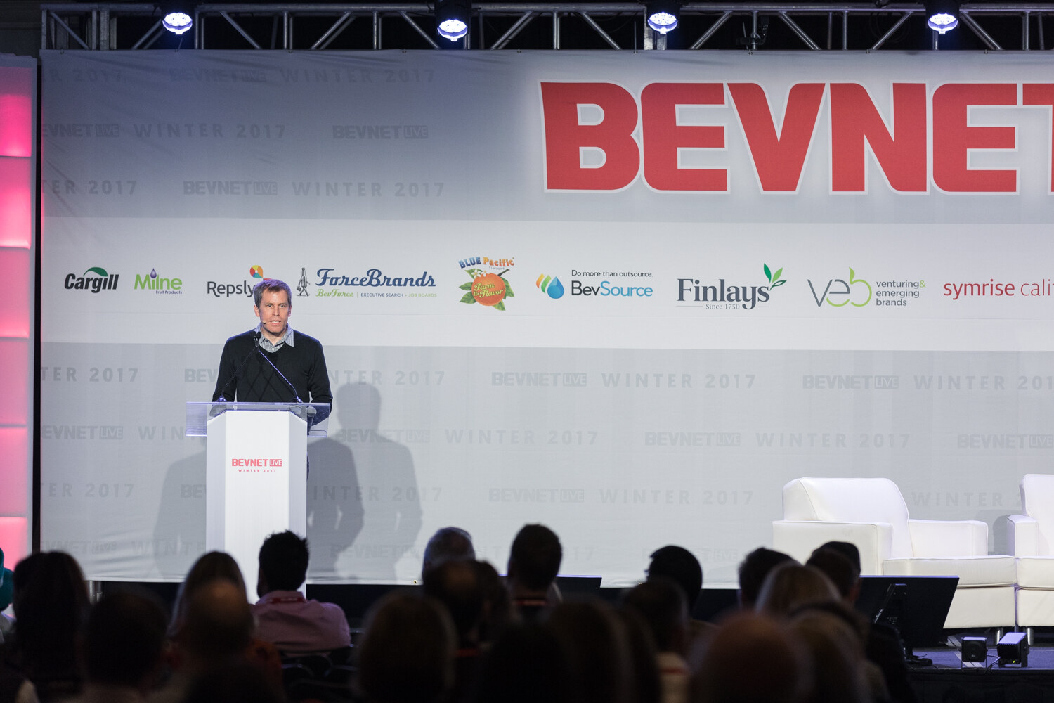 A man in a black sweater speaks at a white lectern in front of a BevNET Live banner with sponsor logos, seen over the heads of the audience.