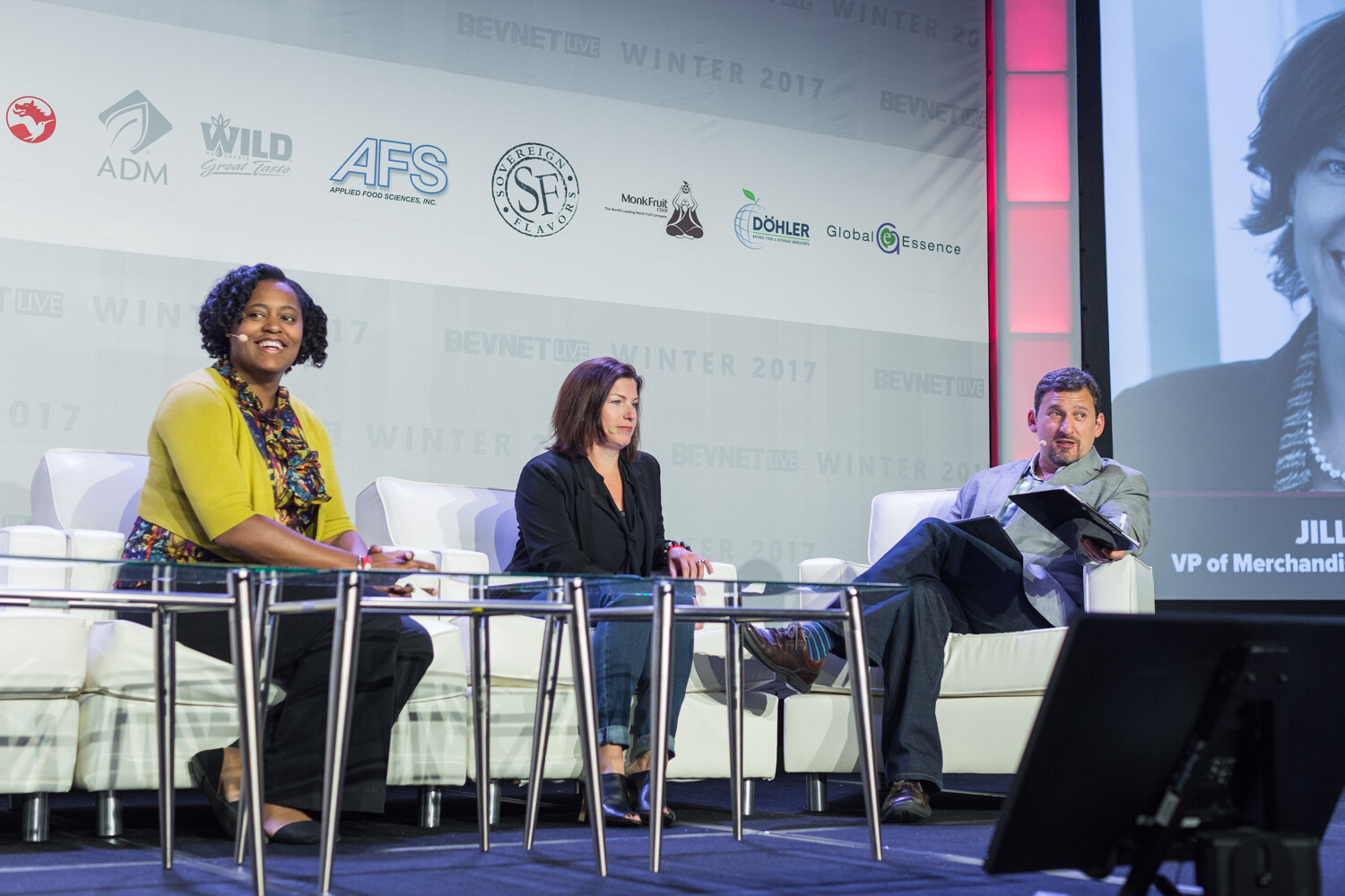 A woman in a yellow cardigan, a woman in a black jacket and a man holding a tablet sit on white sofas on stage, with a screen at right showing a close-up and the caption VP of Merchandising.