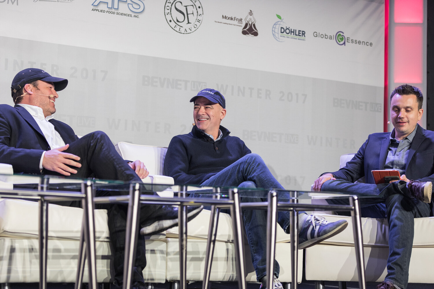Three men on white sofas on stage smile and laugh, two wearing navy caps, with the third holding a red notebook, in front of the sponsor banner.
