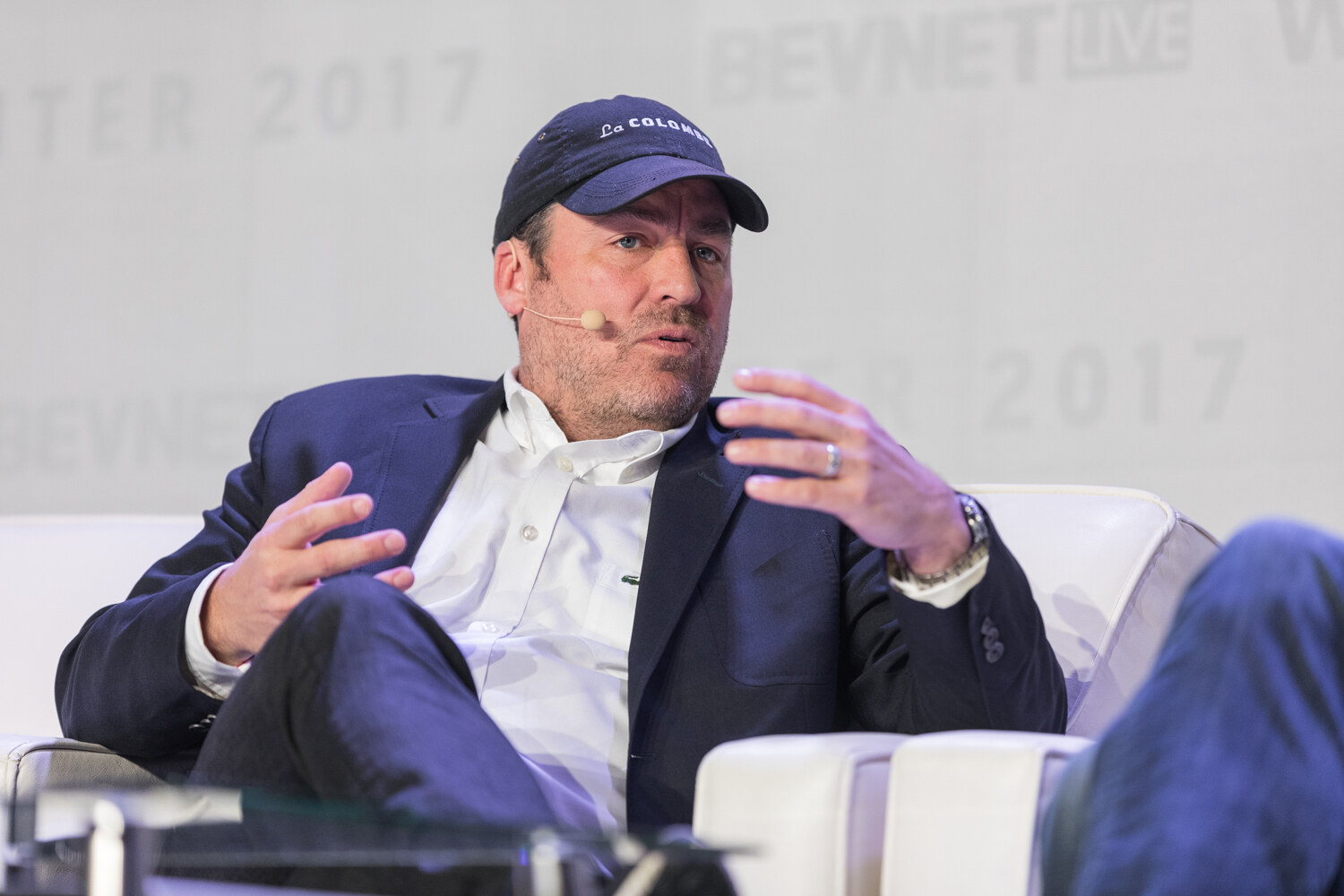 A man in a navy La Colombe baseball cap, navy blazer and white shirt gestures with both hands while seated on a white sofa.